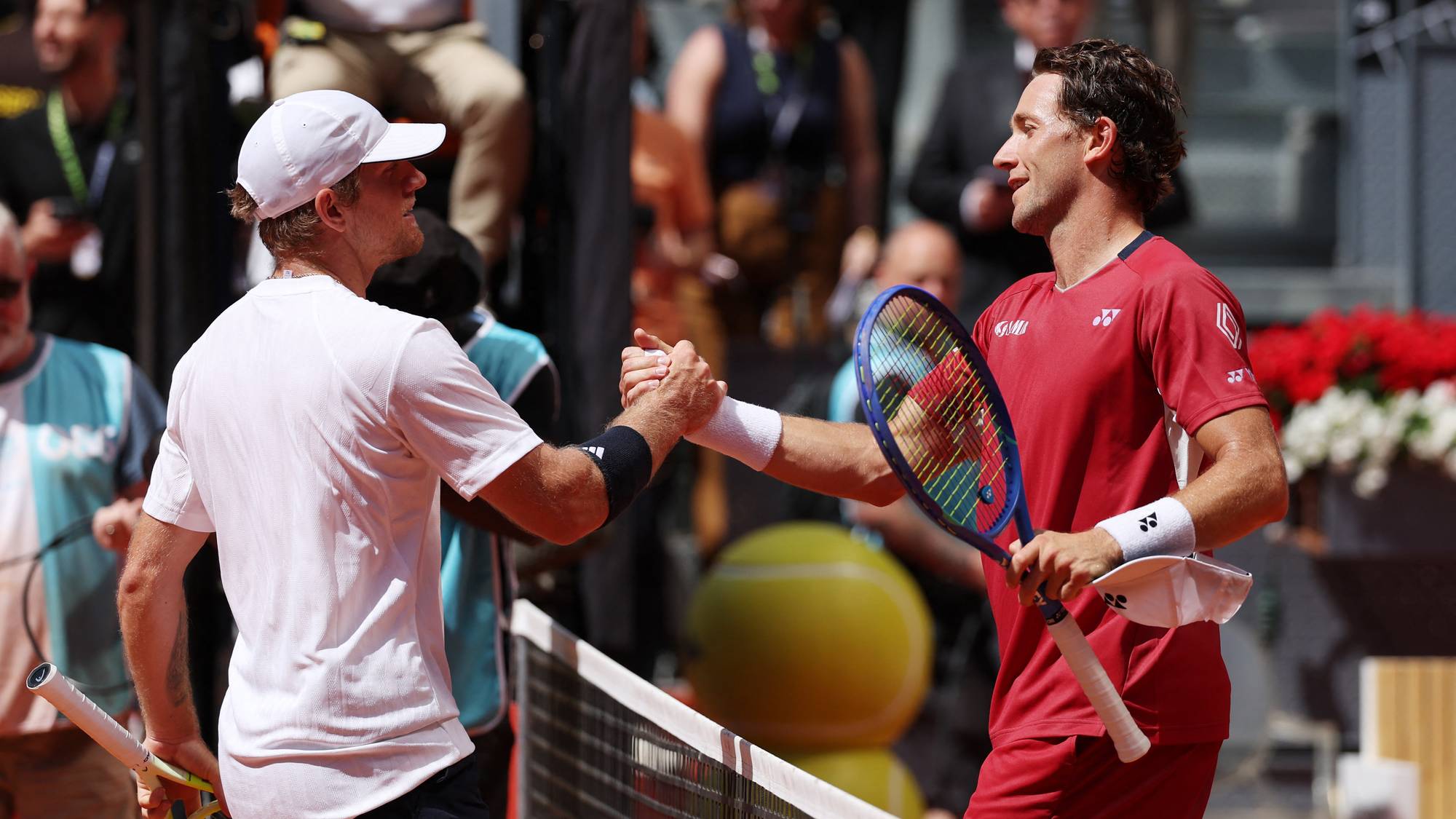 To tennisspillere håndhilser over nettet. Begge er iført sportsantrekk og holder tennisskjermer. Tilskuere er synlige i bakgrunnen. (Bildebeskrivelsen er laget av en KI-tjeneste)