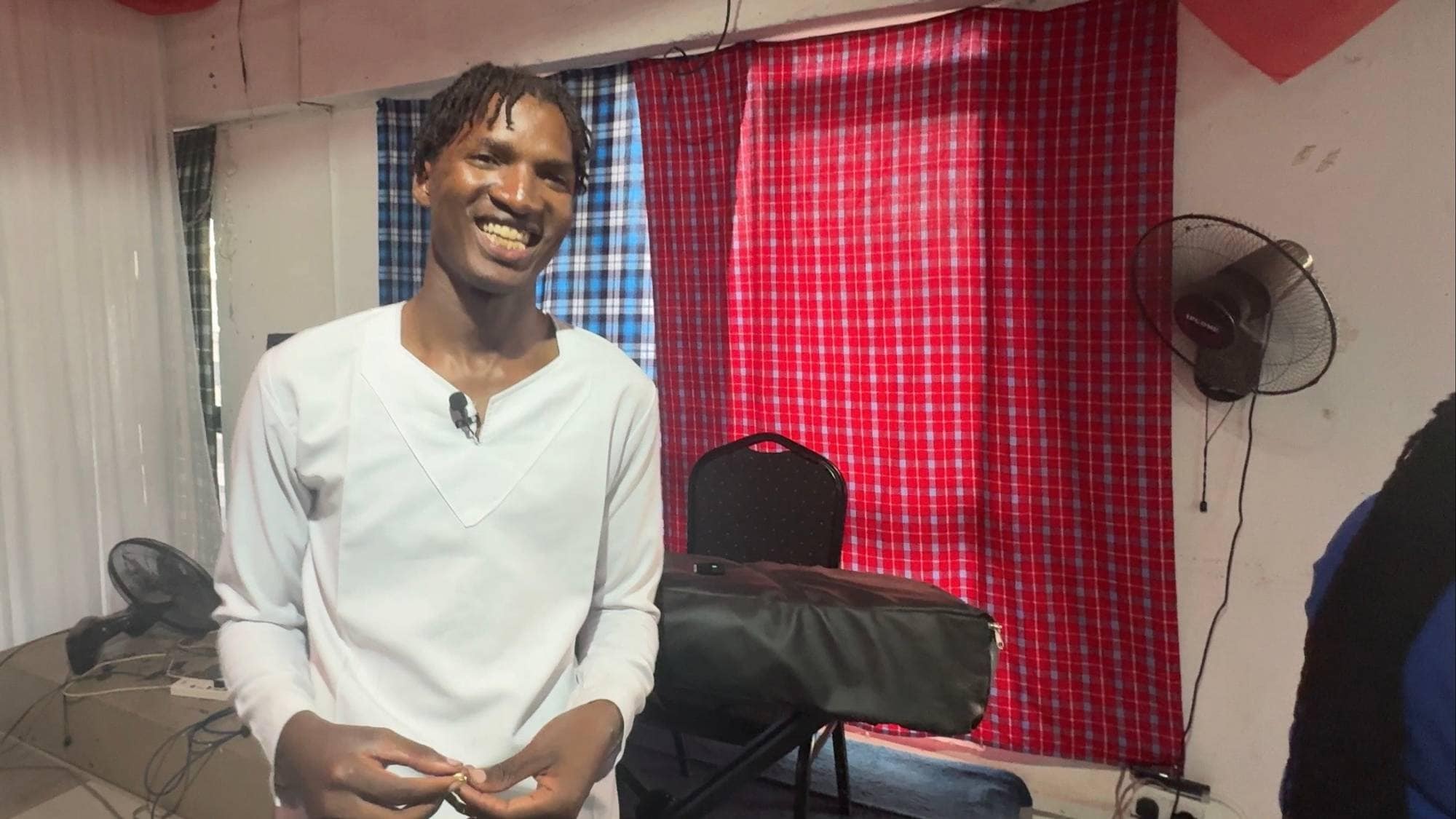 Ein ung kenyansk mann med mørk hud og eit stort smil ser mot kamera. Han ser snill ut, kledd i ei kvit skjorte. Han står i eit rom med store fargerike tepper framfor vindauga.