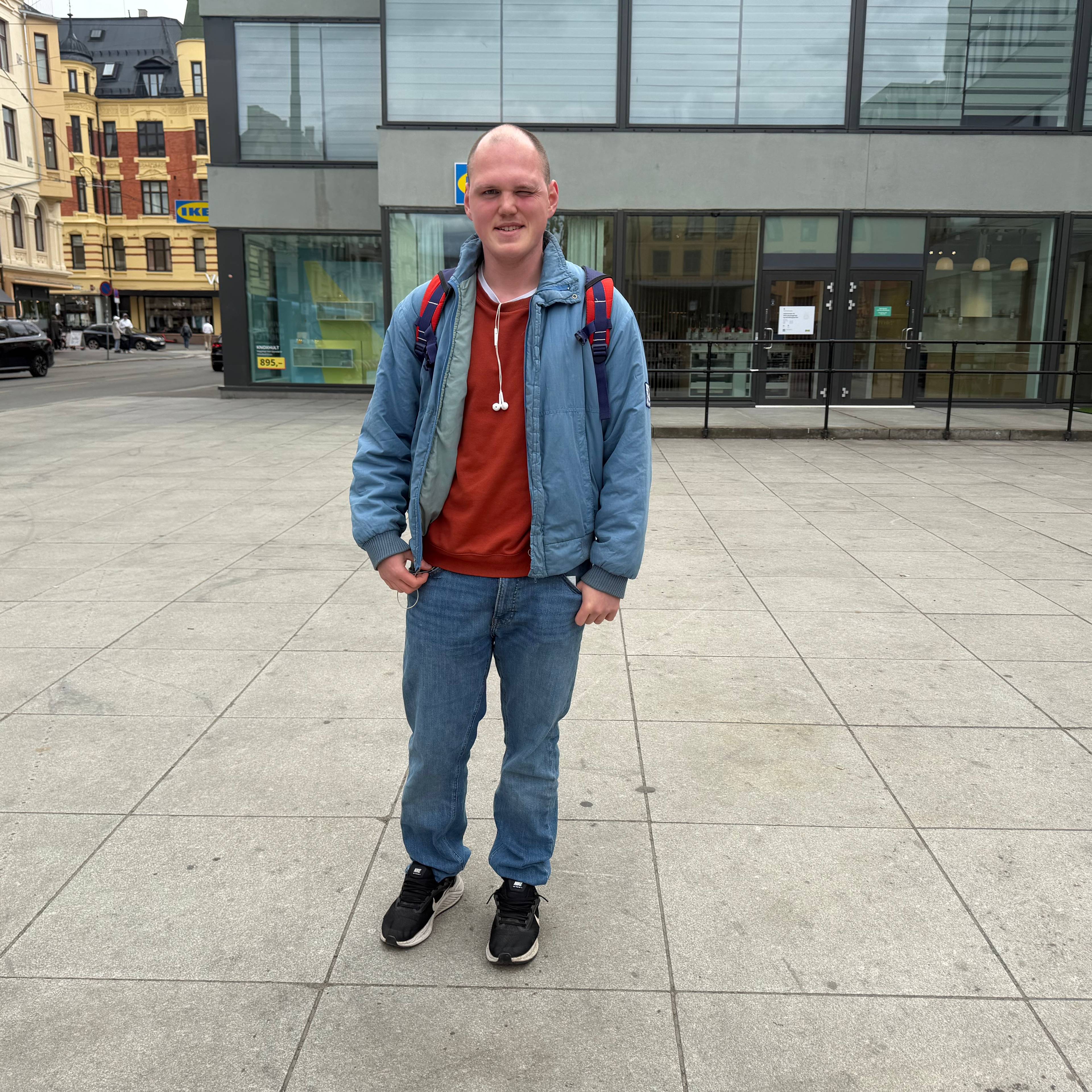 En mann står på et brosteinsbelagt torg foran en bygning med store glassflater. Mannen har på seg en blå jakke, oransje genser og jeans. I bakgrunnen sees bygninger og en gate med biler. (Bildebeskrivelsen er laget av en KI-tjeneste)