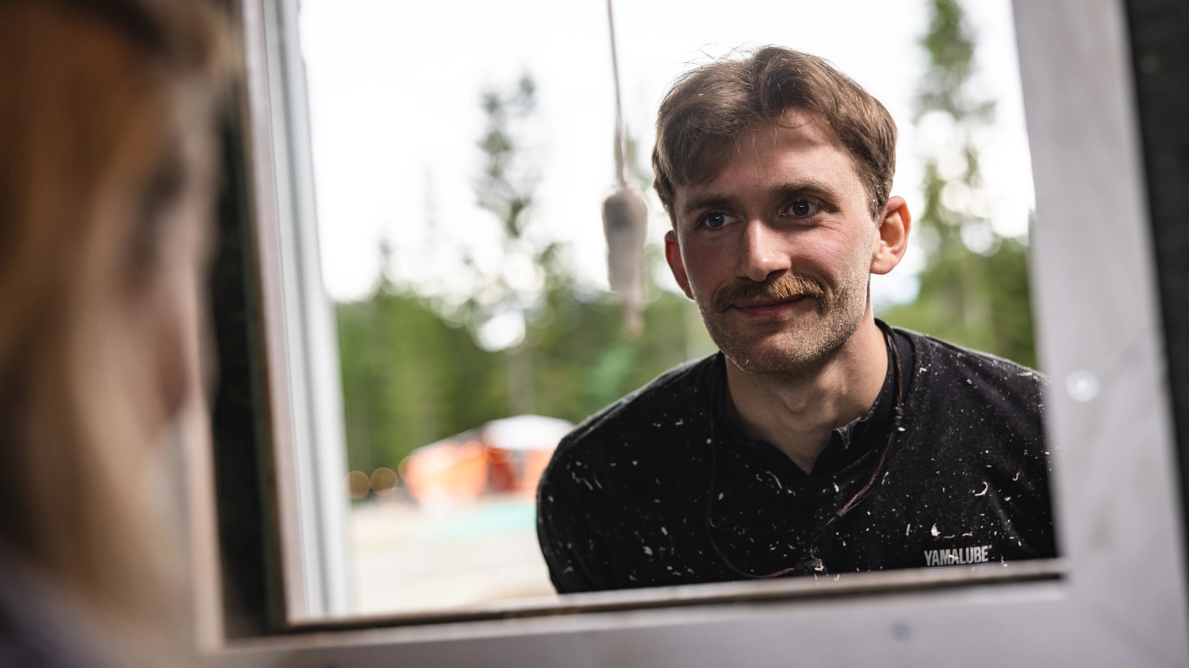 Sivert og Tora snakker sammen gjennom en åpning i en vegg. Vi ser så vidt håret og øret til Tora, og hele ansiktet til Sivert - som smiler