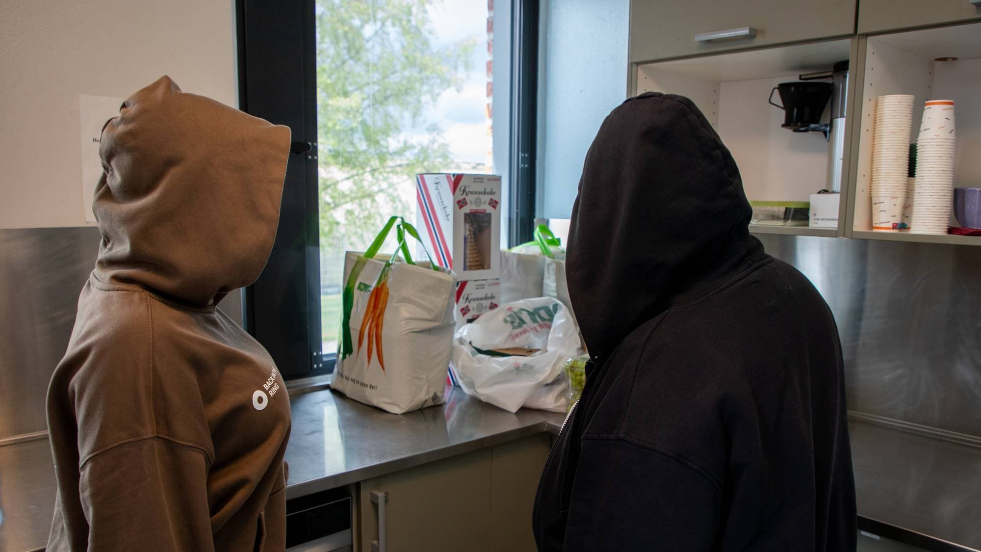 To kvinner med store hettegensre står med ryggen til kameraet. Foran dem er en kjøkkenbenk med blant annet kransekake og plastglass synlig.