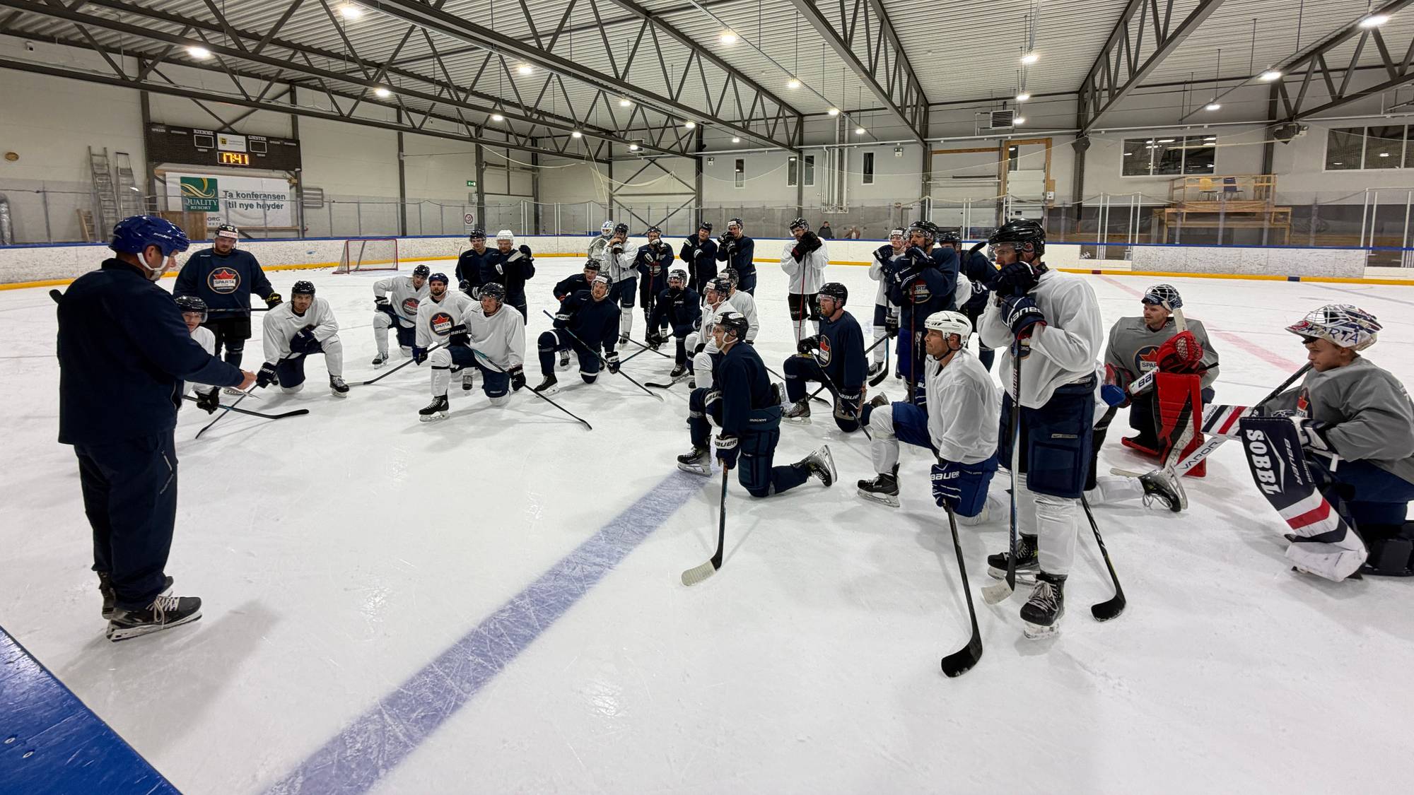 Ishockeylag samlet på isen med trener som gir instruksjoner under en treningsøkt. (Bildebeskrivelsen er laget av en KI-tjeneste)