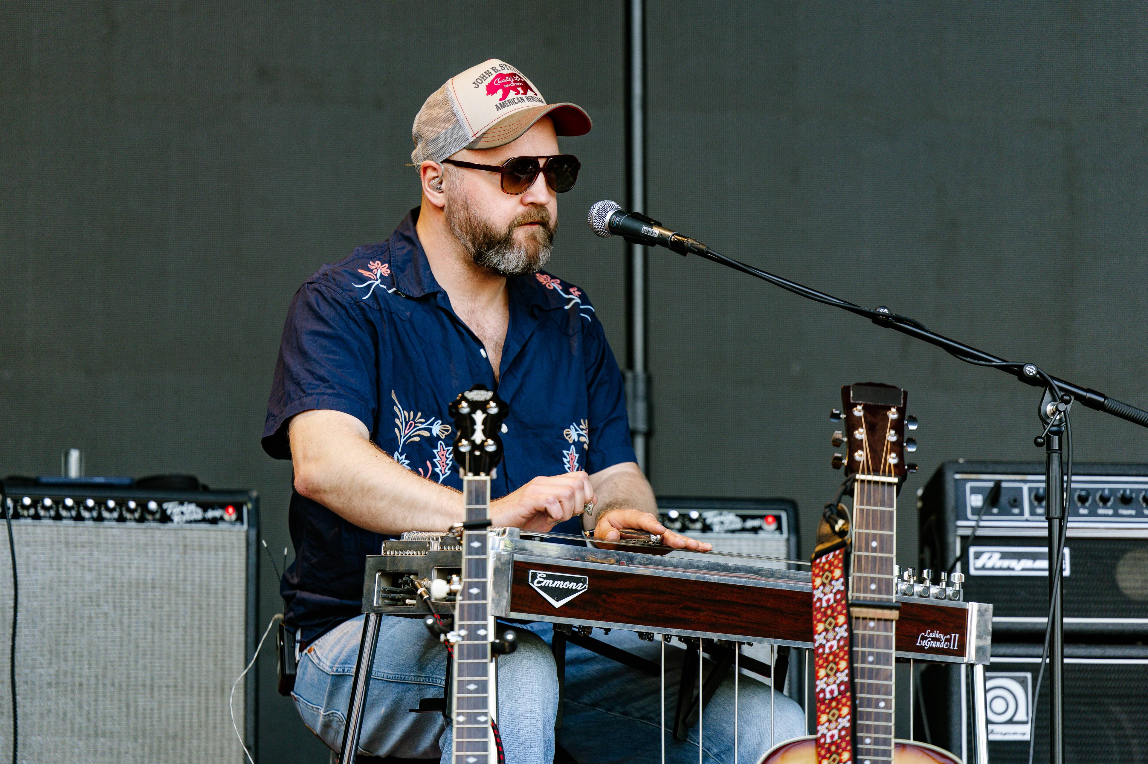 En mann sitter og spiller steelgitar på en scene. Han har på seg solbriller, caps og en mørkeblå skjorte. Det står gitarforsterkere bak ham. (Bildebeskrivelsen er laget av en KI-tjeneste)