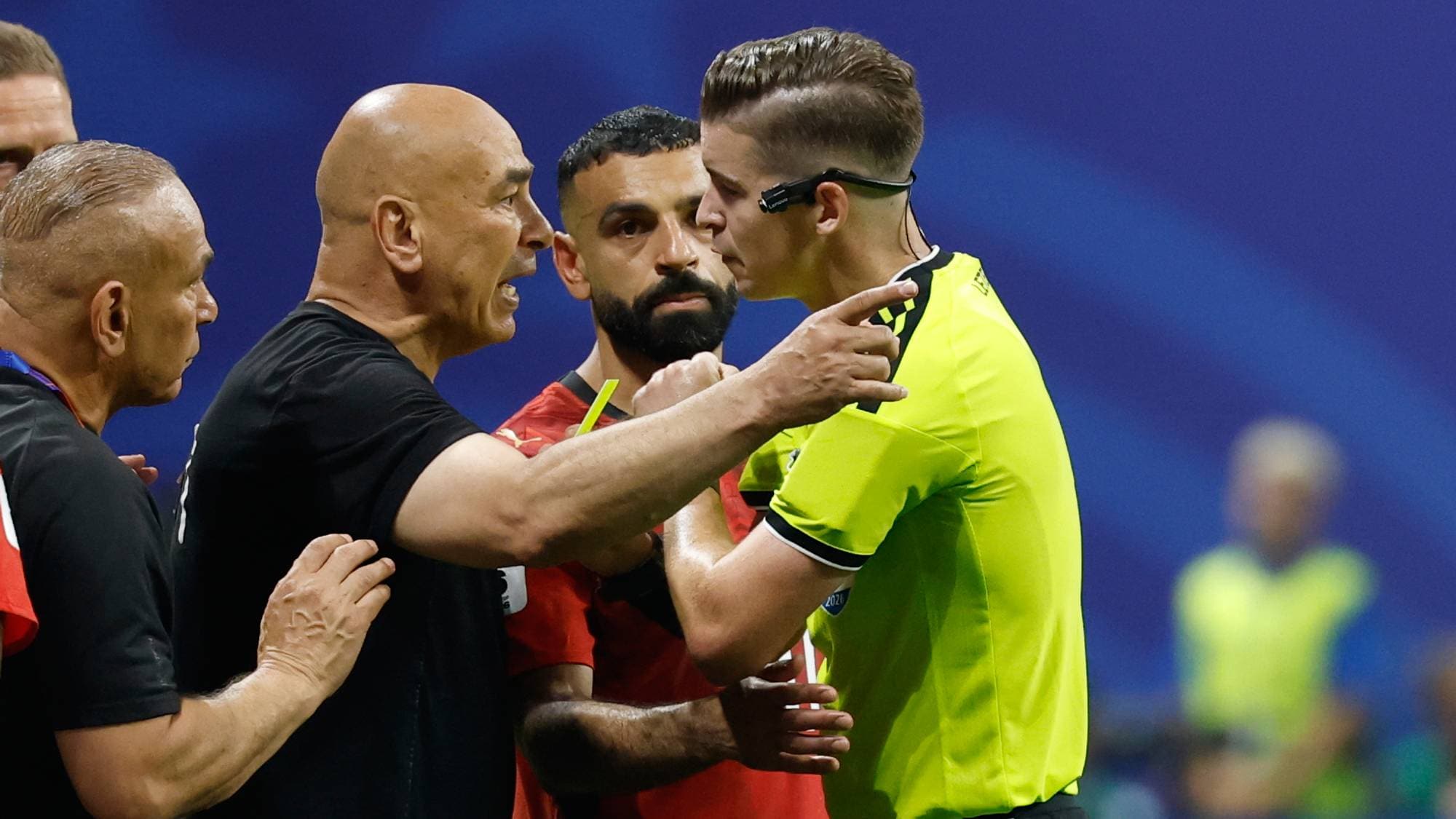 Fotballtrener diskuterer med dommeren mens en spiller ser på under en kamp. (Bildebeskrivelsen er laget av en KI-tjeneste)