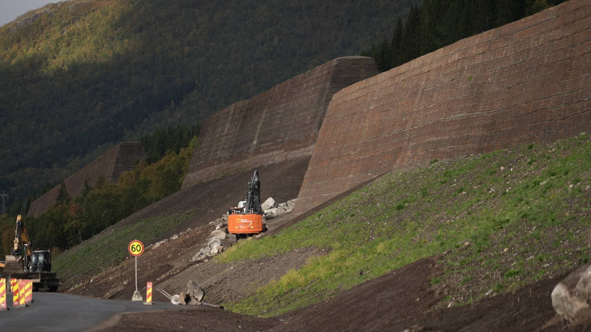 Anleggsmaskiner jobber ved en stor skredvoll langs en vei i et fjellområde. (Bildebeskrivelsen er laget av en KI-tjeneste)
