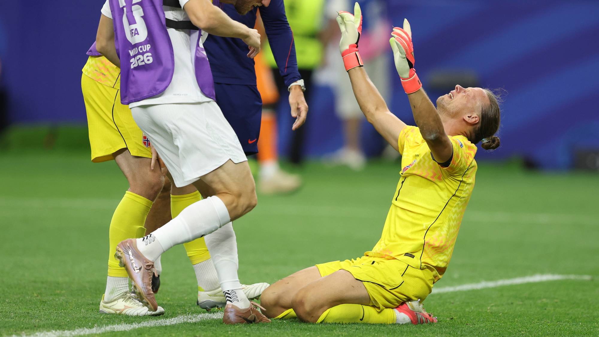 Norsk keeper sitter på gresset med armene i været, mens lagkamerater kommer bort for å trøste. (Bildebeskrivelsen er laget av en KI-tjeneste)