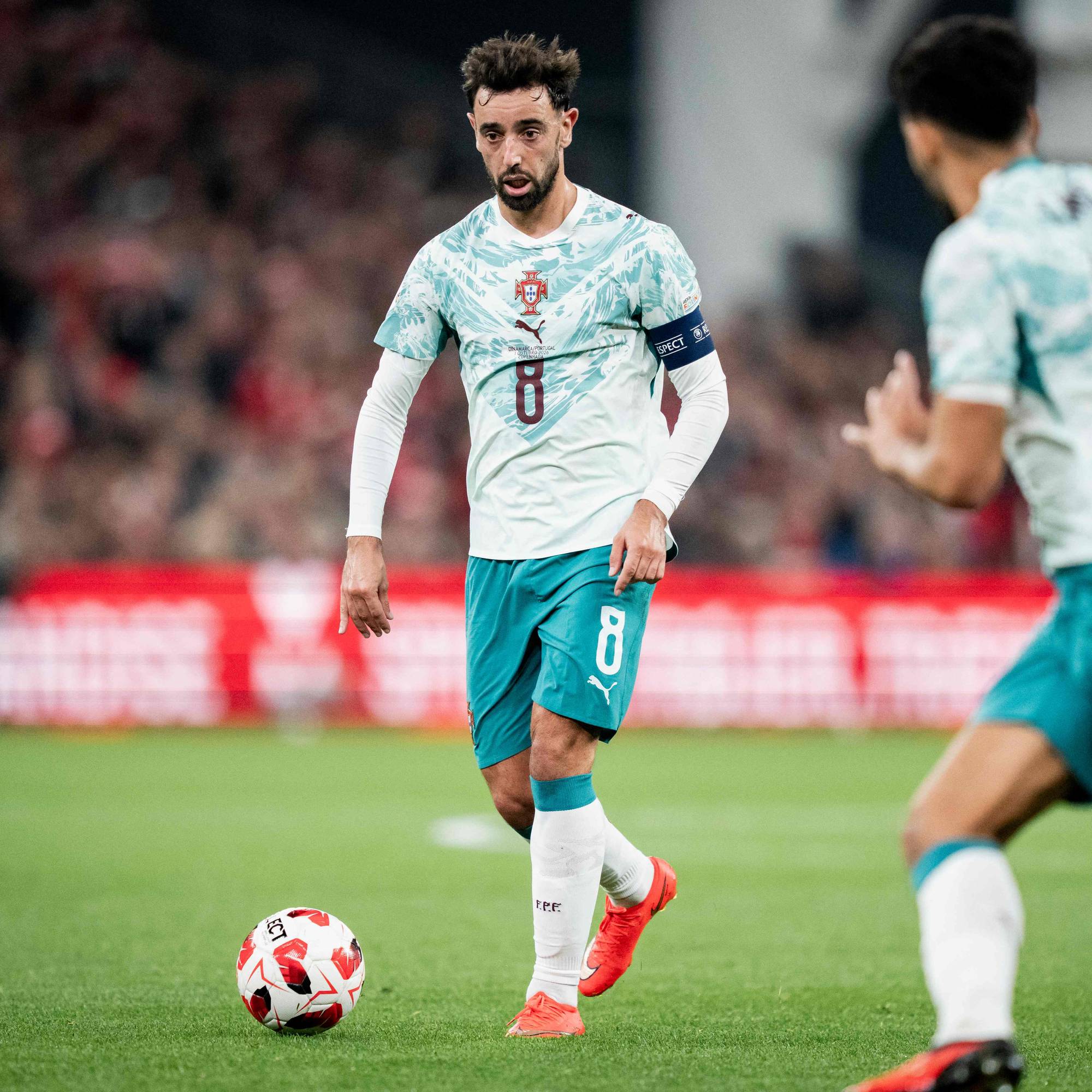 Bruno Fernandes fører ballen på fotballbanen under en kamp for Portugal. Lagkamerat i bakgrunnen. (Bildebeskrivelsen er laget av en KI-tjeneste)