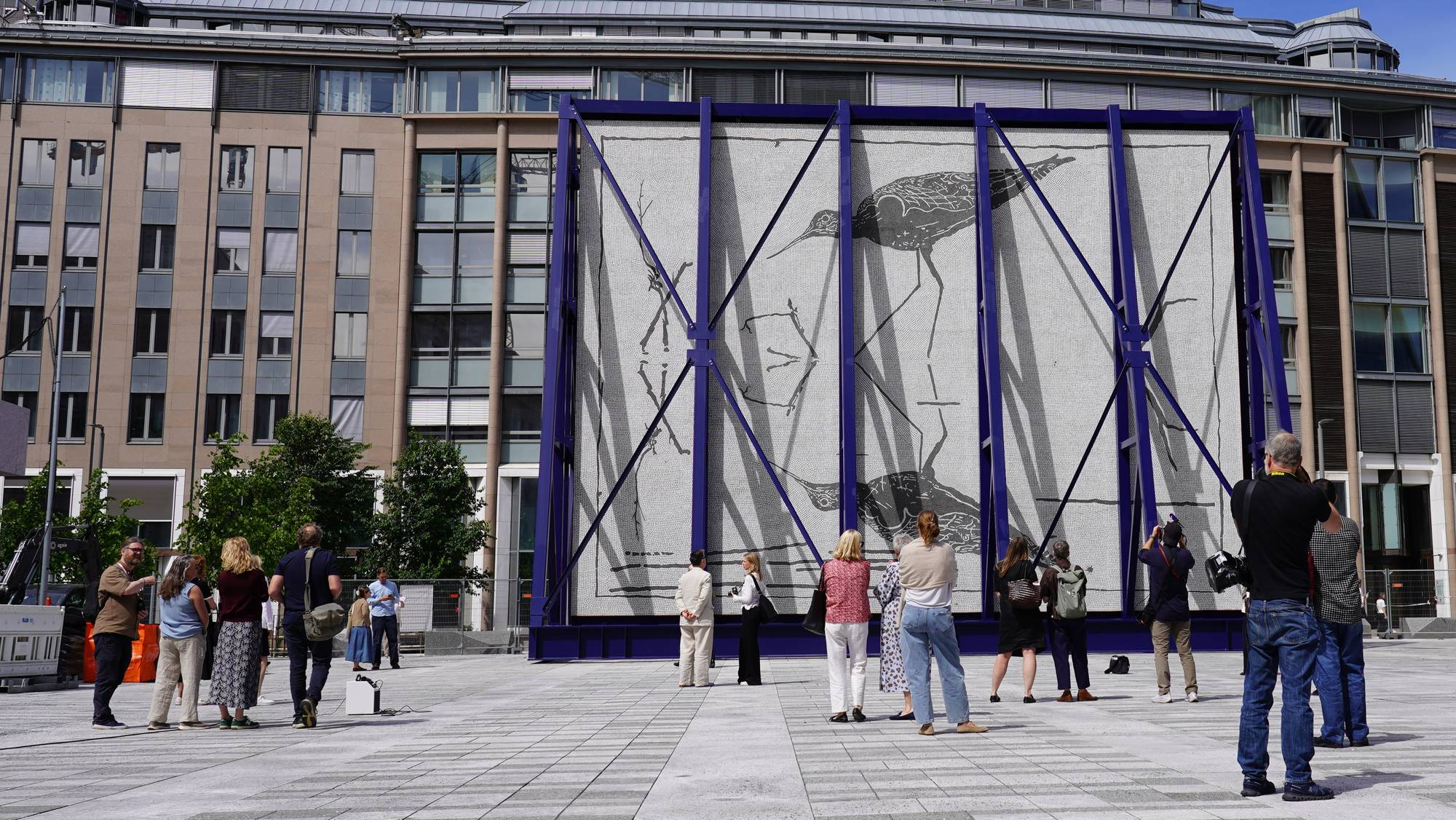 Folk ser på en stor installasjon med et bilde av to fugler. Installasjonen er dekket av tusenvis av små steiner. Folk står på et flislagt torg med moderne bygninger i bakgrunnen. (Bildebeskrivelsen er laget av en KI-tjeneste)