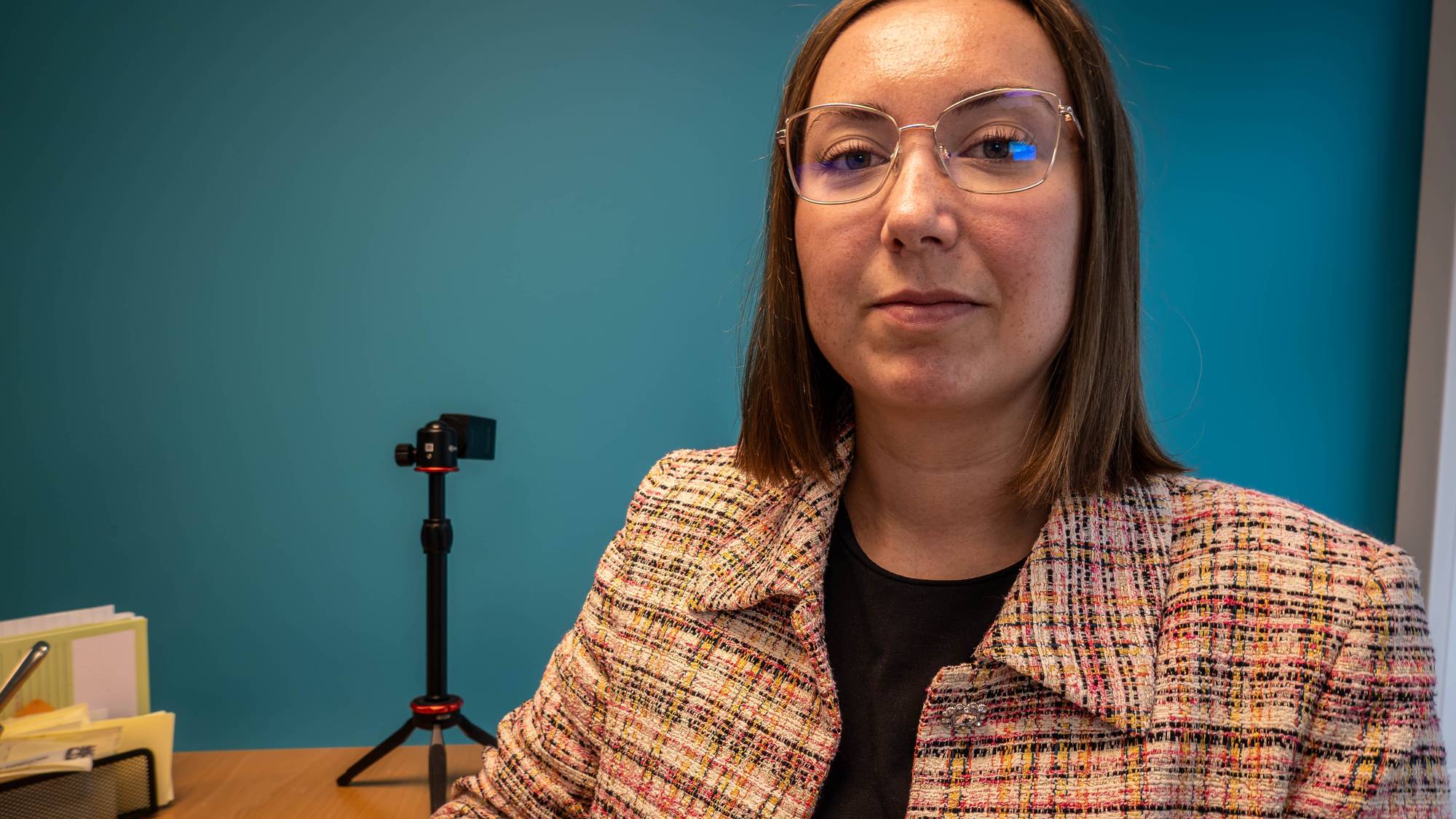 Kvinne med briller foran en blå vegg ved et kontorpult med et lite kamerastativ. (Bildebeskrivelsen er laget av en KI-tjeneste)