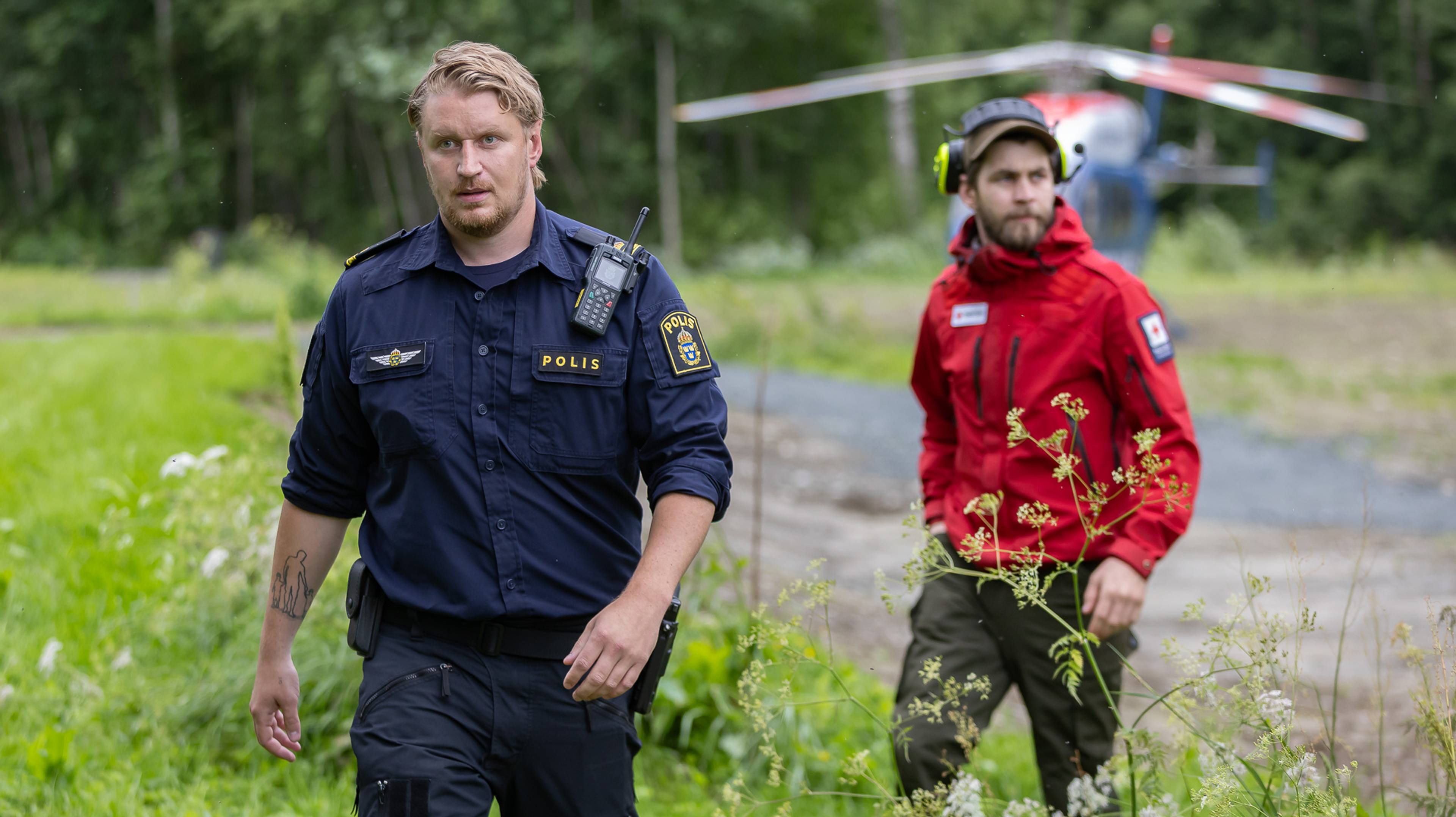 En mann i svensk politiuniform kommer gående mot fotografen. Bak ham ser vi en annen mann fra Røde Kors. I bakgrunnen står et helikopter