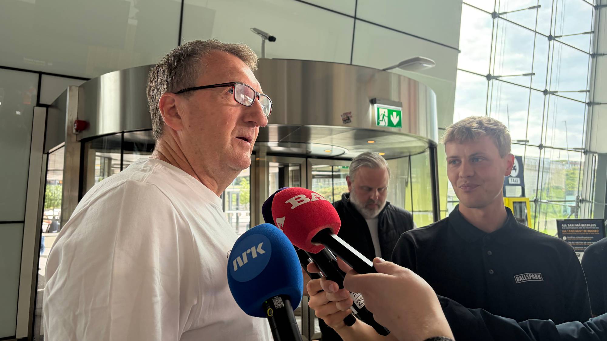 Mann i hvit t-skjorte blir intervjuet av pressefolk med mikrofoner i et moderne terminalbygg. (Bildebeskrivelsen er laget av en KI-tjeneste)