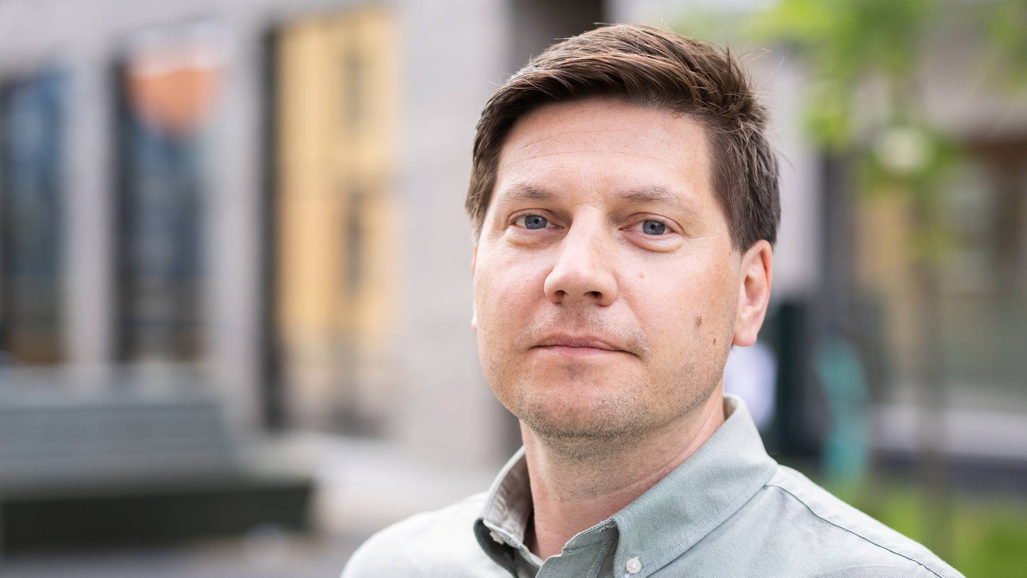 En mann i grå skjorte som smiler og ser inn i kamera, med et bygg og hage i bakgrunnen.