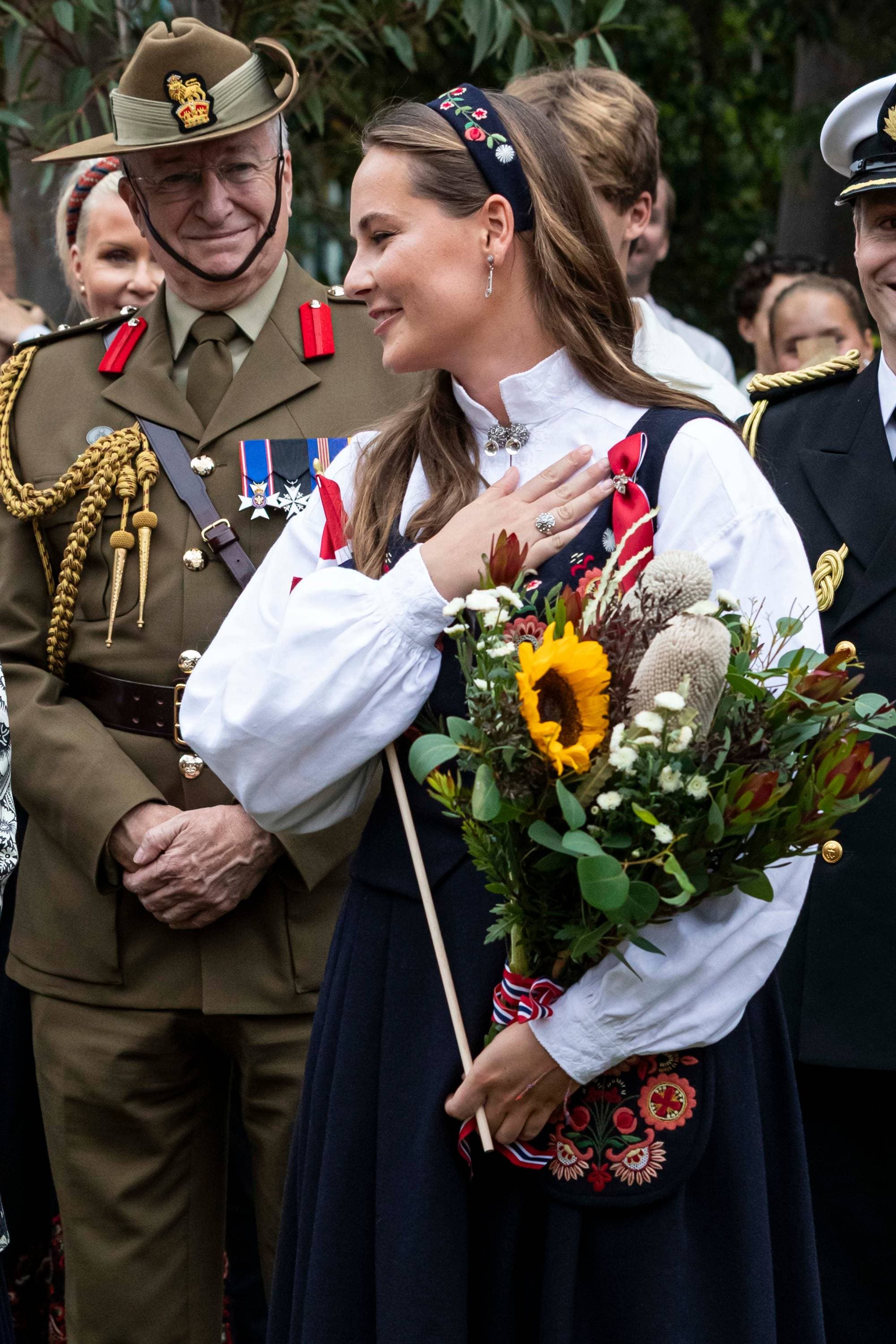 Prinsesse Ingrid Alexandra i svart bunad med blomsterbukett med gul solsikke. Ved siden står en mann i grønn uniform og bredbremmet grønn hatt brettet opp på siden. 