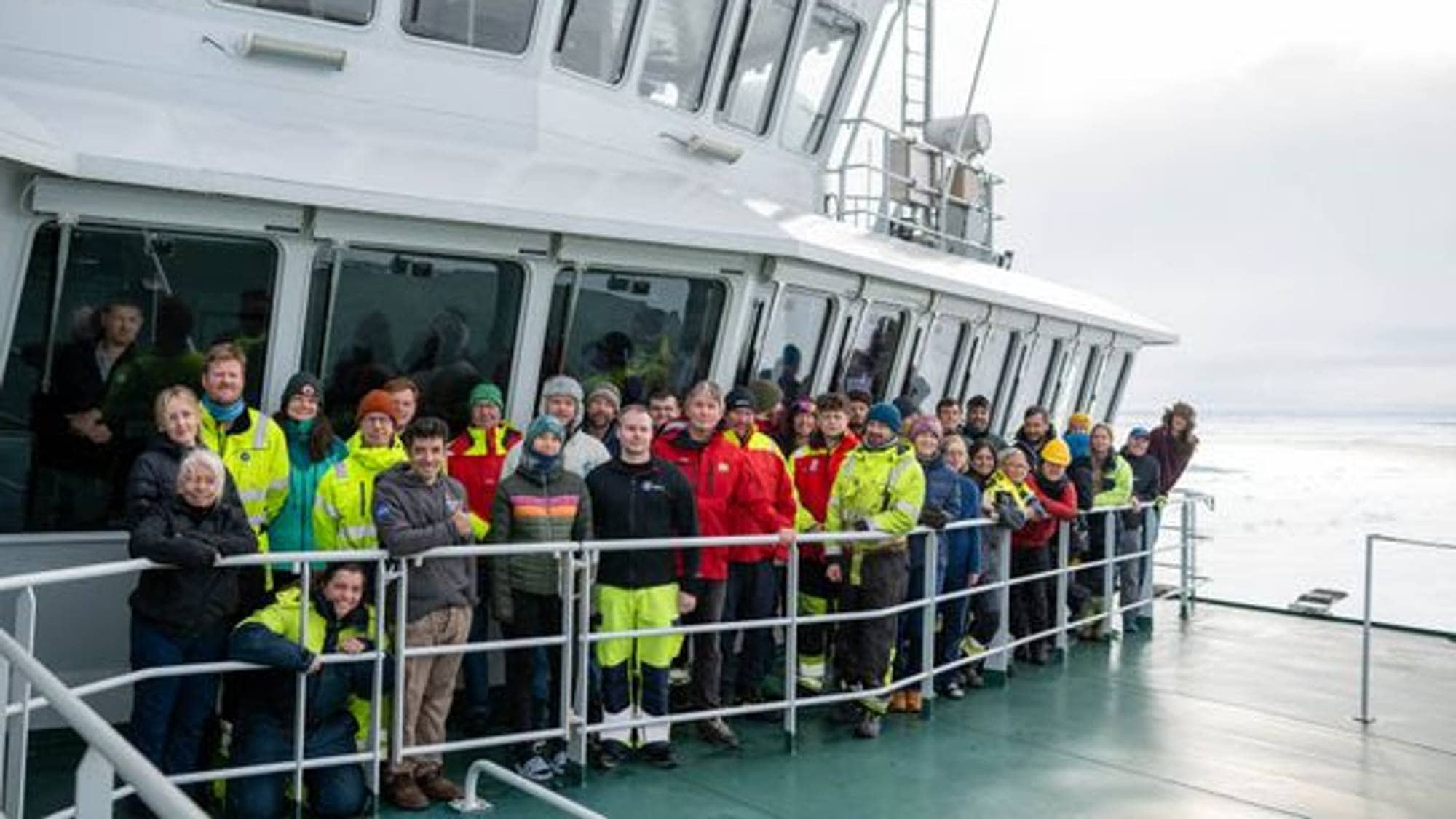 En stor gruppe mennesker står oppstilt på dekk på et skip i arktiske omgivelser. 