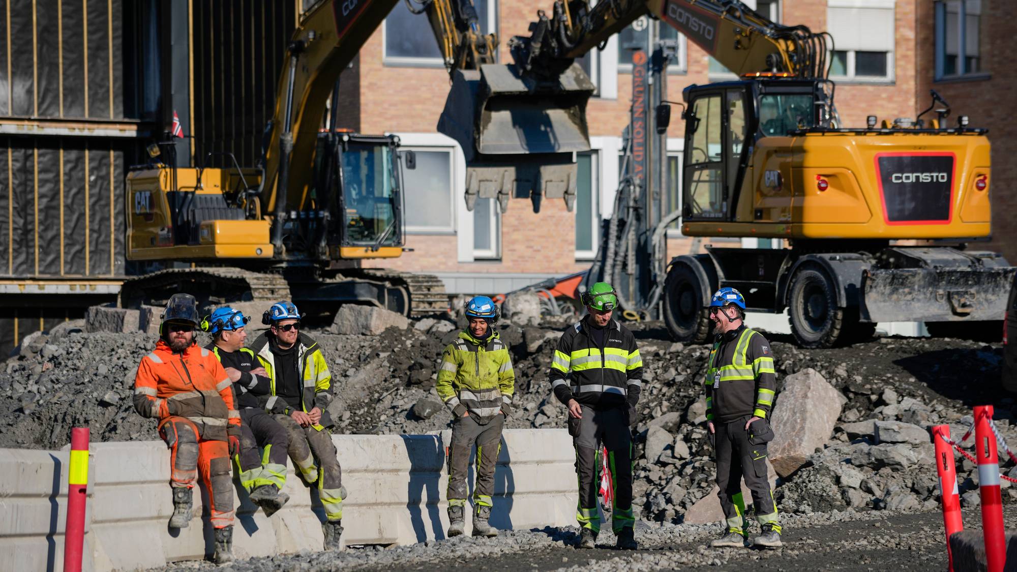 Fem anleggsarbeidere står og sitter på en grå betongblokk foran to gule gravemaskiner på en byggeplass. Tre av mennene sitter på blokken, mens to står. De er alle iført verneklær og hjelm. I bakgrunnen er det en mursteinsbygning. Solen skinner og kaster skygger på bakken. (Bildebeskrivelsen er laget av en KI-tjeneste)