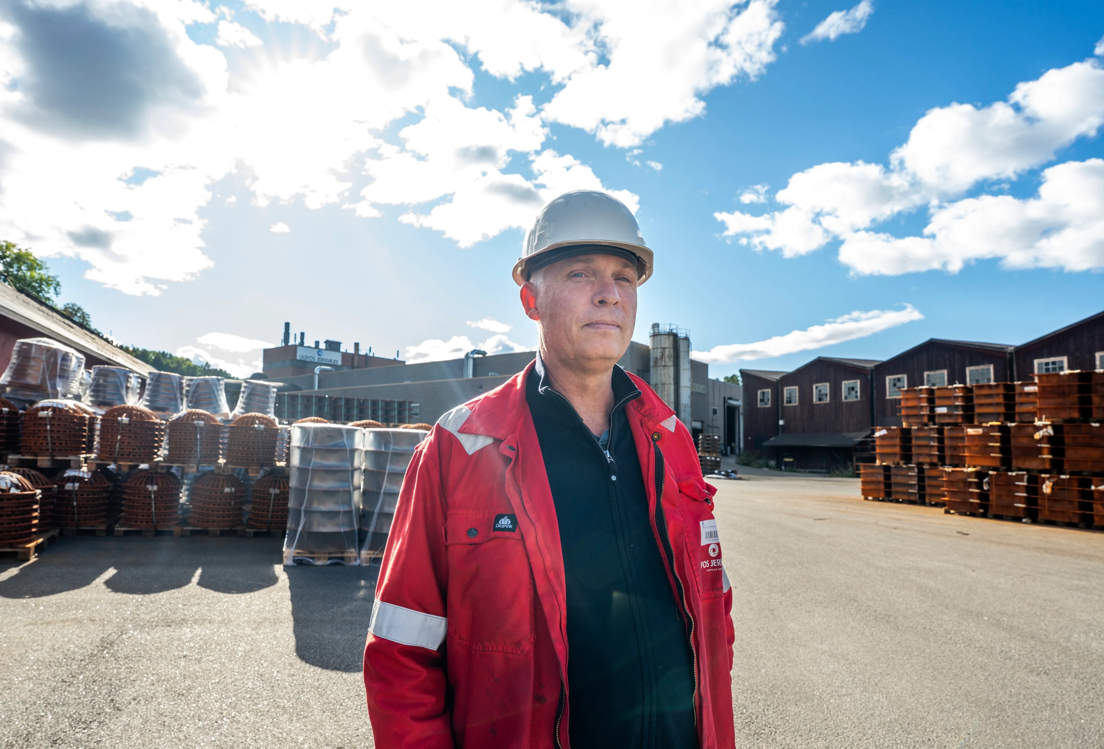 En mann i vernehjelm og rød jakke står på et industriområde med paller av støpejern. (Bildebeskrivelsen er laget av en KI-tjeneste)