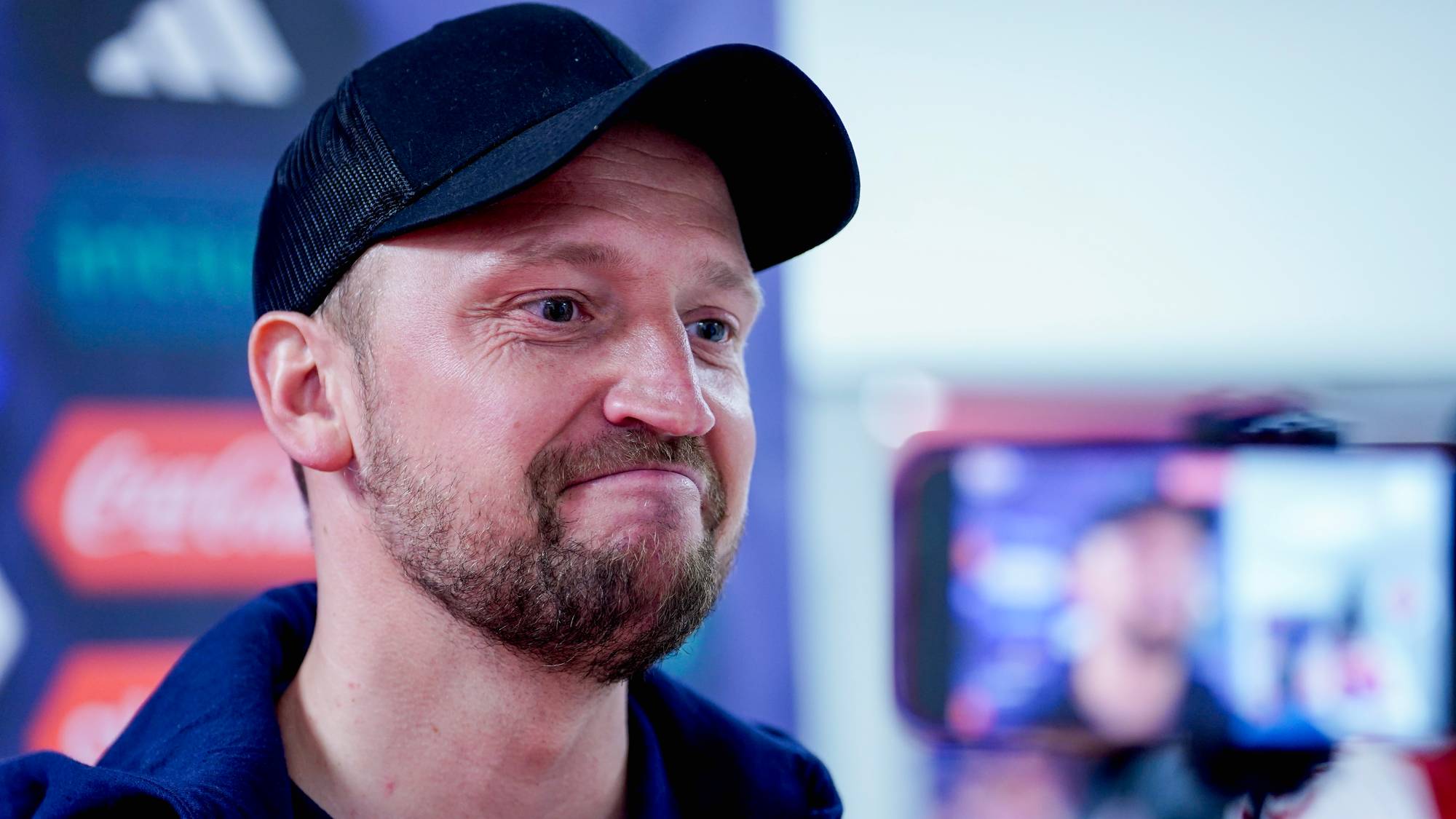 Johannes Moesgaard, trener for KFUM, smiler. Han har på seg svart caps og en blå jakke over en mørk genser.