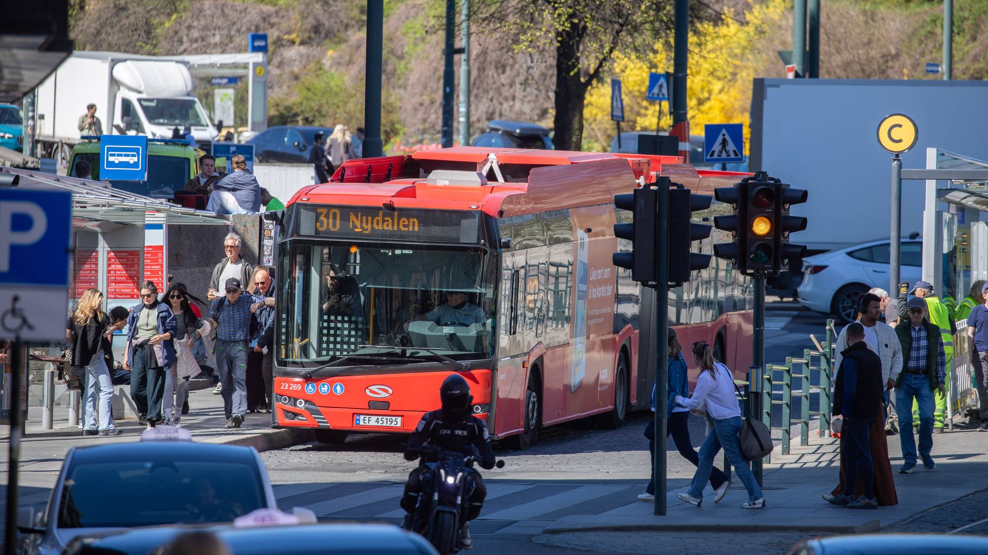 En buss med nummer 30 og destinasjon Nydalen står ved et busstopp. Flere personer venter på bussen. En motorsyklist kjører forbi. (Bildebeskrivelsen er laget av en KI-tjeneste)