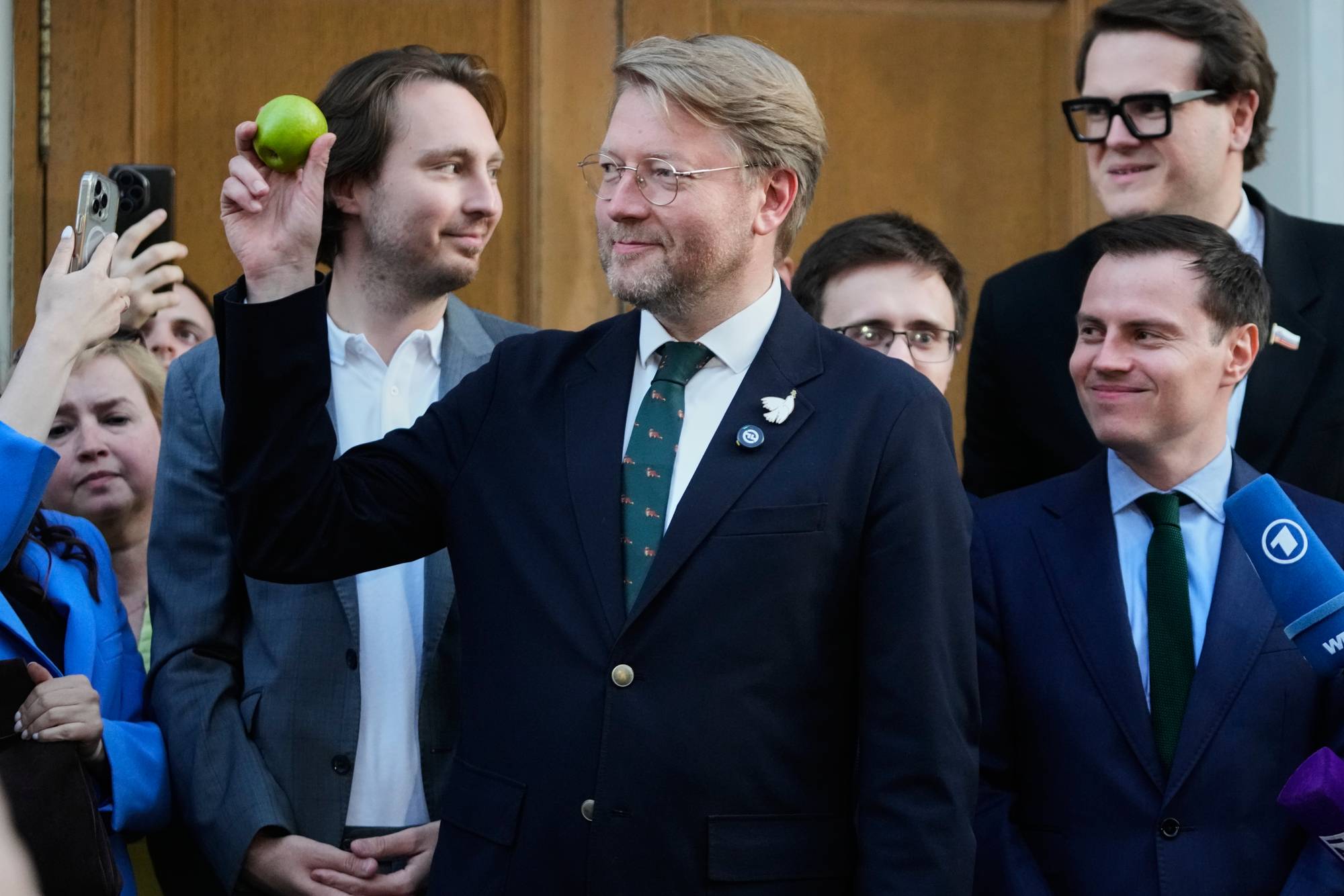 Partileder Nikolai Rybakov står foran retten, Han er blond og har runde briller, og er kledd i dress. Han holder et eple som han vise r fram til publikum. Bak han står flere menn kledd i dress. 