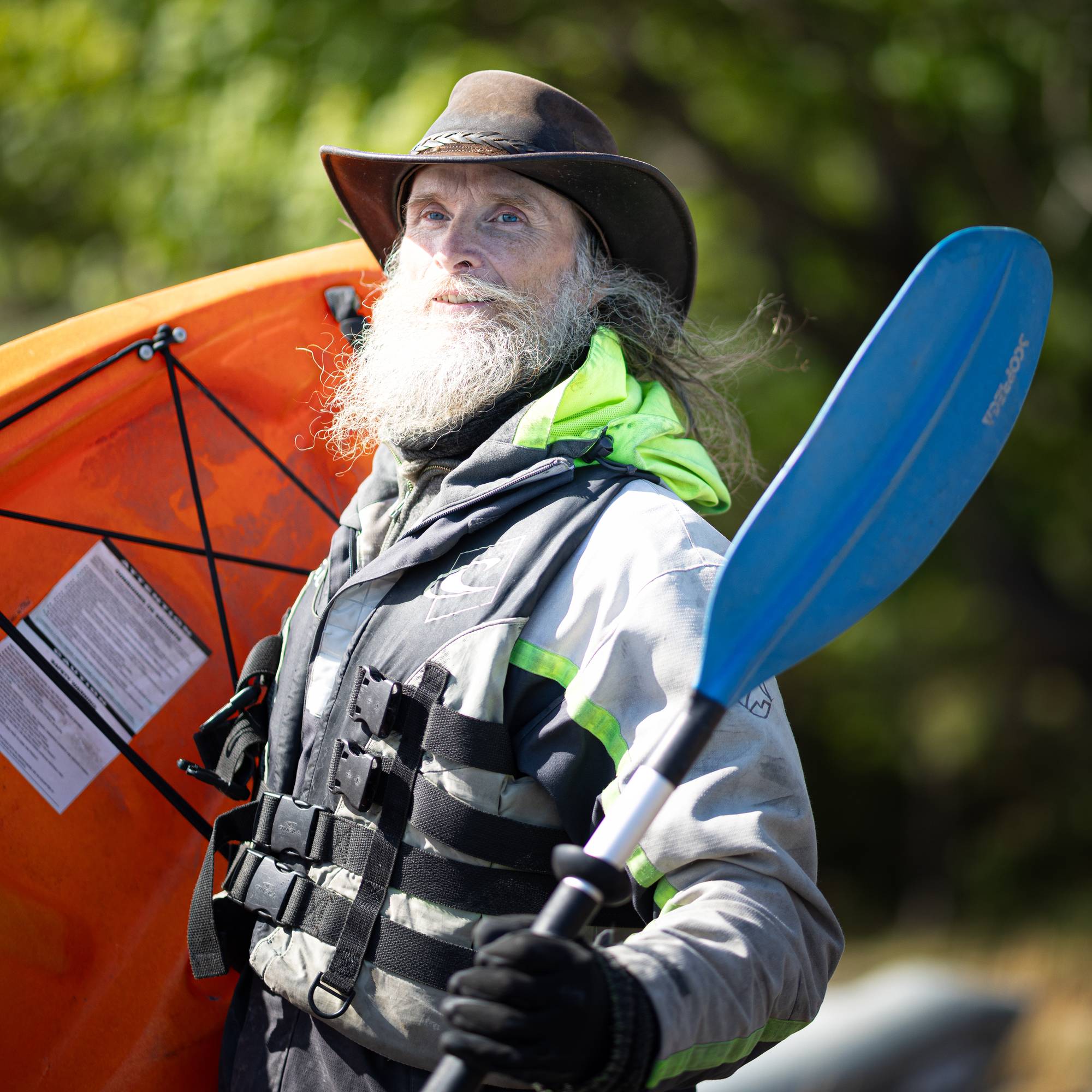 En mann med langt, grått skjegg og cowboyhatt. Han har på seg redningsvest og holder en kaijak og en året i hånden. Han står utendørs og ser mot kamera.