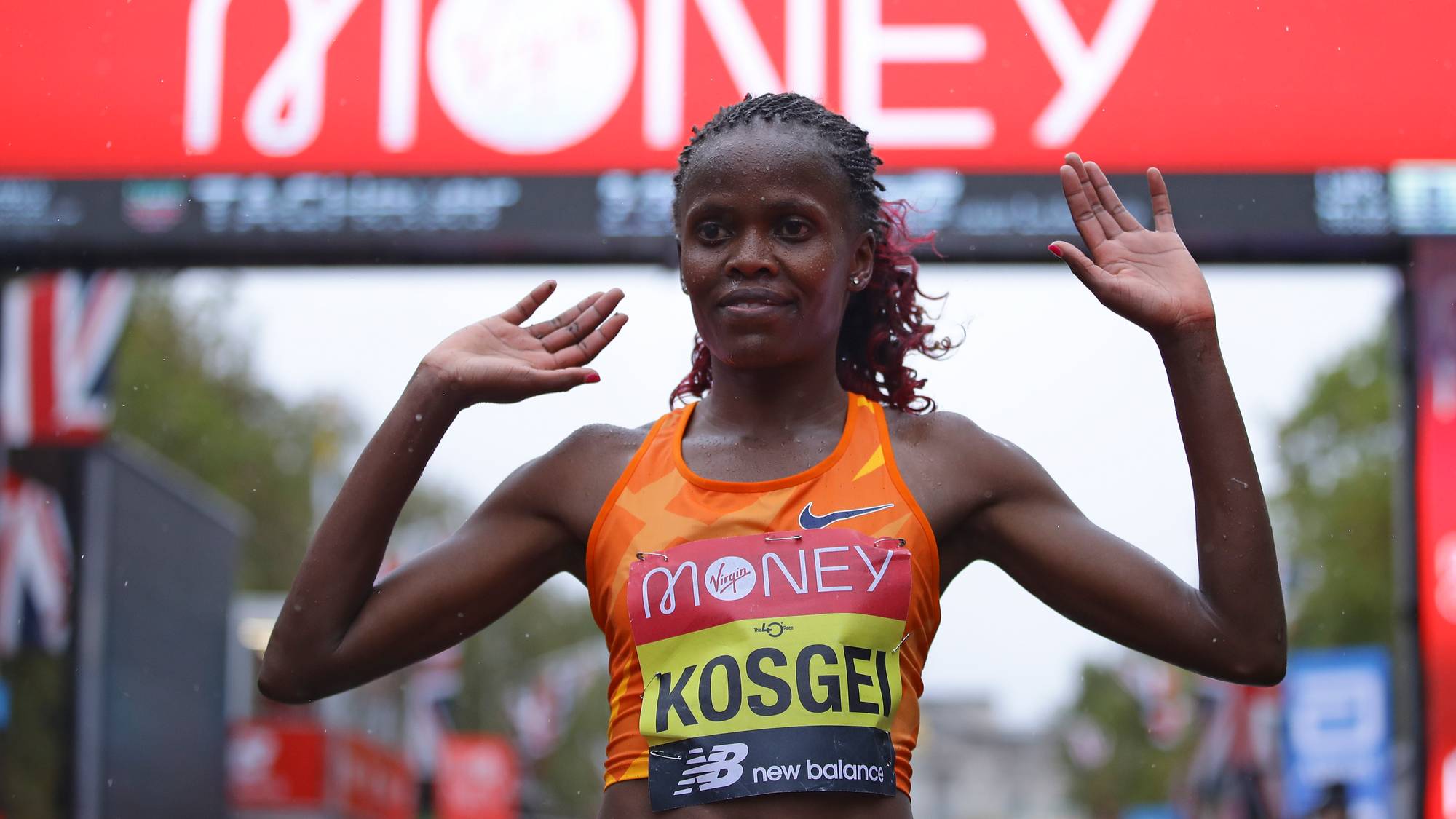 En kvinne i en oransje løpevest og med navnet KOSGEI på brystet hever hendene. Bak henne er en rød bue med teksten "MONEY". (Bildebeskrivelsen er laget av en KI-tjeneste)