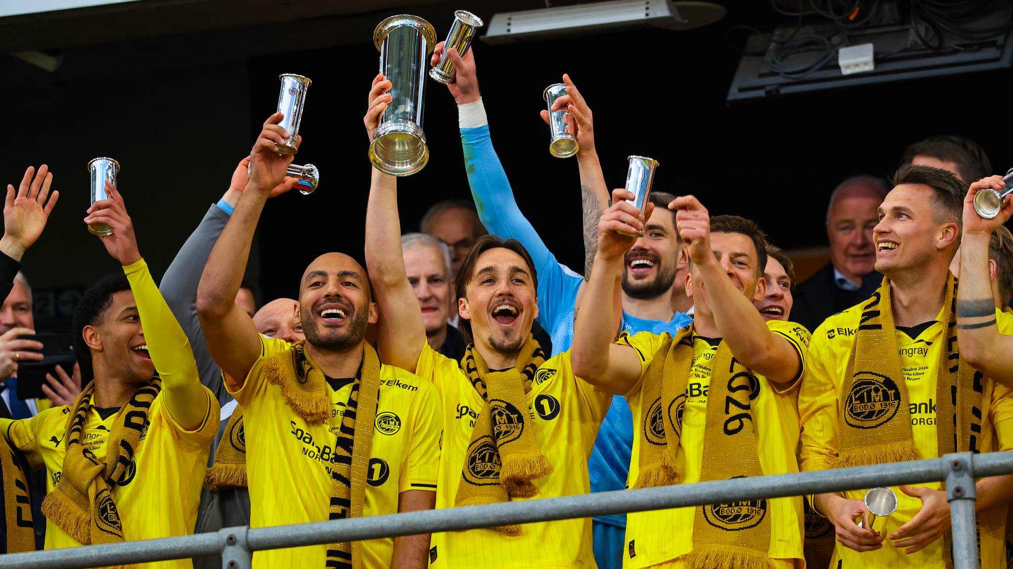 Fotballspillere i gule drakter feirer med pokaler. Flere spillere holder opp pokaler og smiler. De har gule skjerf rundt halsen. Bildet er tatt utendørs under en seremoni. (Bildebeskrivelsen er laget av en KI-tjeneste)