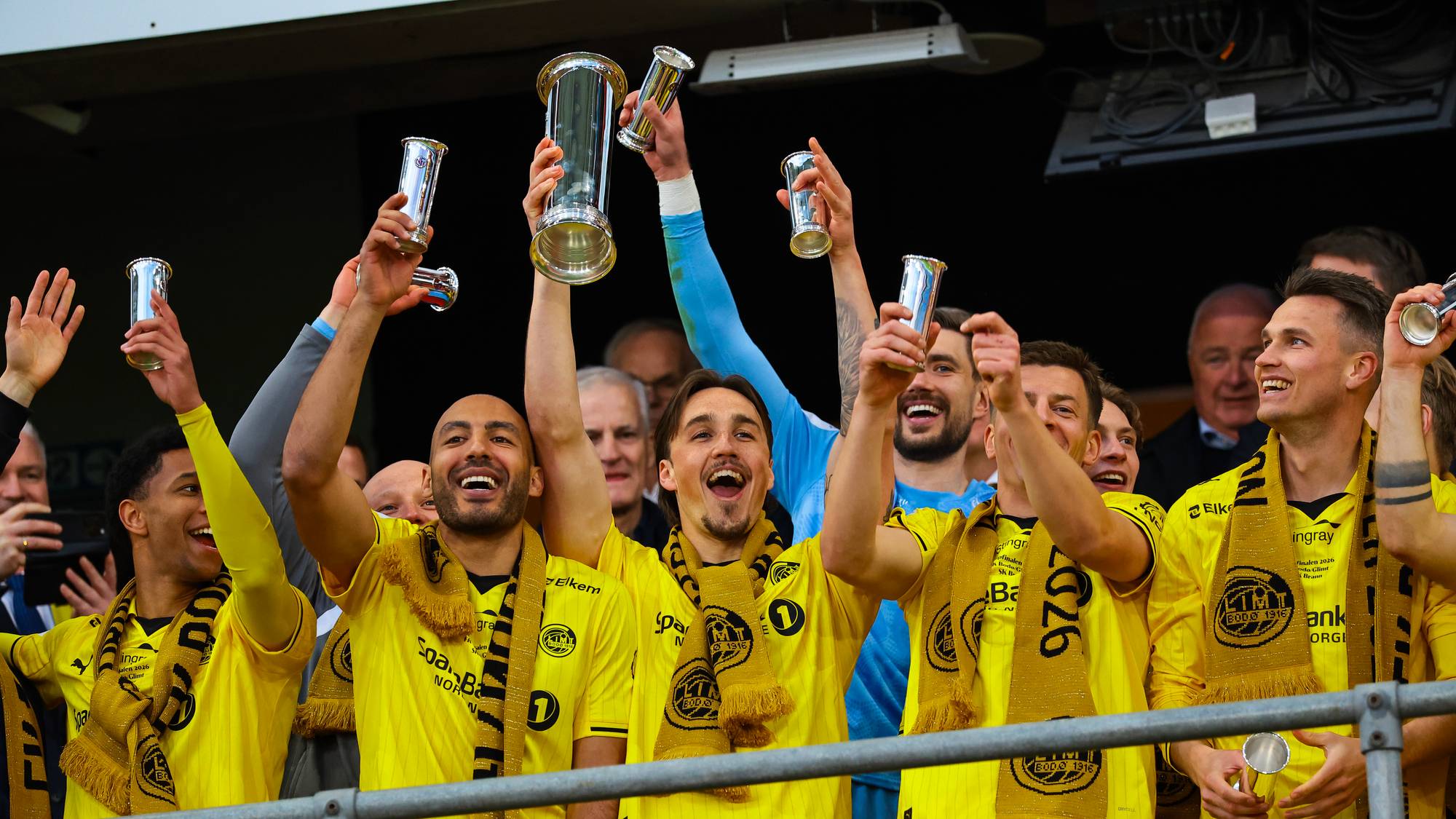 Fotballspillere i gule drakter feirer med pokaler. Flere spillere holder opp pokaler og smiler. De har gule skjerf rundt halsen. Bildet er tatt utendørs under en seremoni. (Bildebeskrivelsen er laget av en KI-tjeneste)
