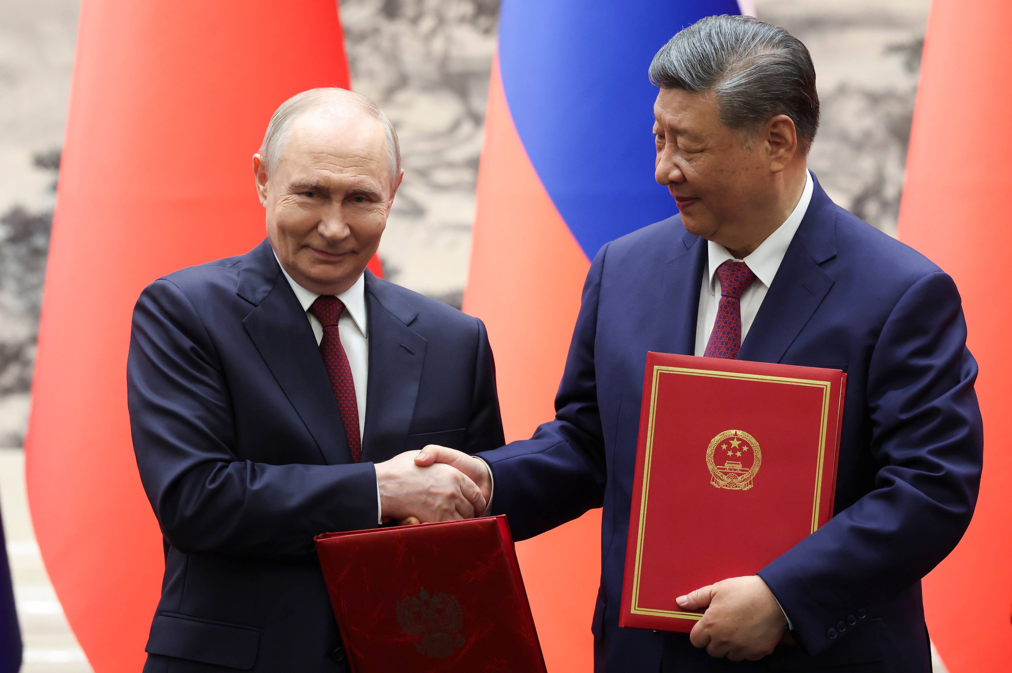 Russlands president Vladimir Putin og Kinas president Xi Jinping håndhilser. De holder røde mapper med dokumenter. Bak dem er store røde og blå flagg. (Bildebeskrivelsen er laget av en KI-tjeneste)