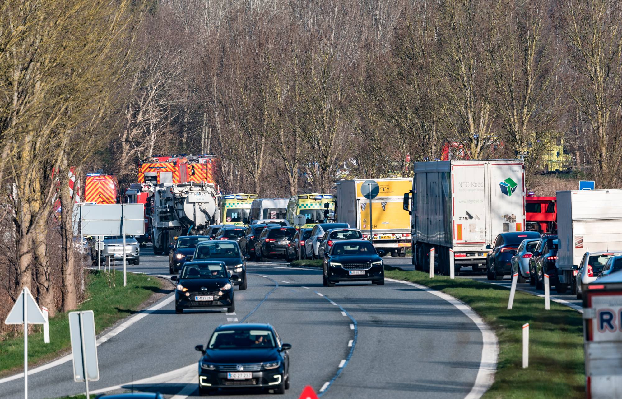 Trafikkhendelse med mange utrykningskjøretøy på en vei. Det er en lang kø av personbiler og lastebiler som står stille. Blant utrykningskjøretøyene er det brannbiler og ambulanser. (Bildebeskrivelsen er laget av en KI-tjeneste)