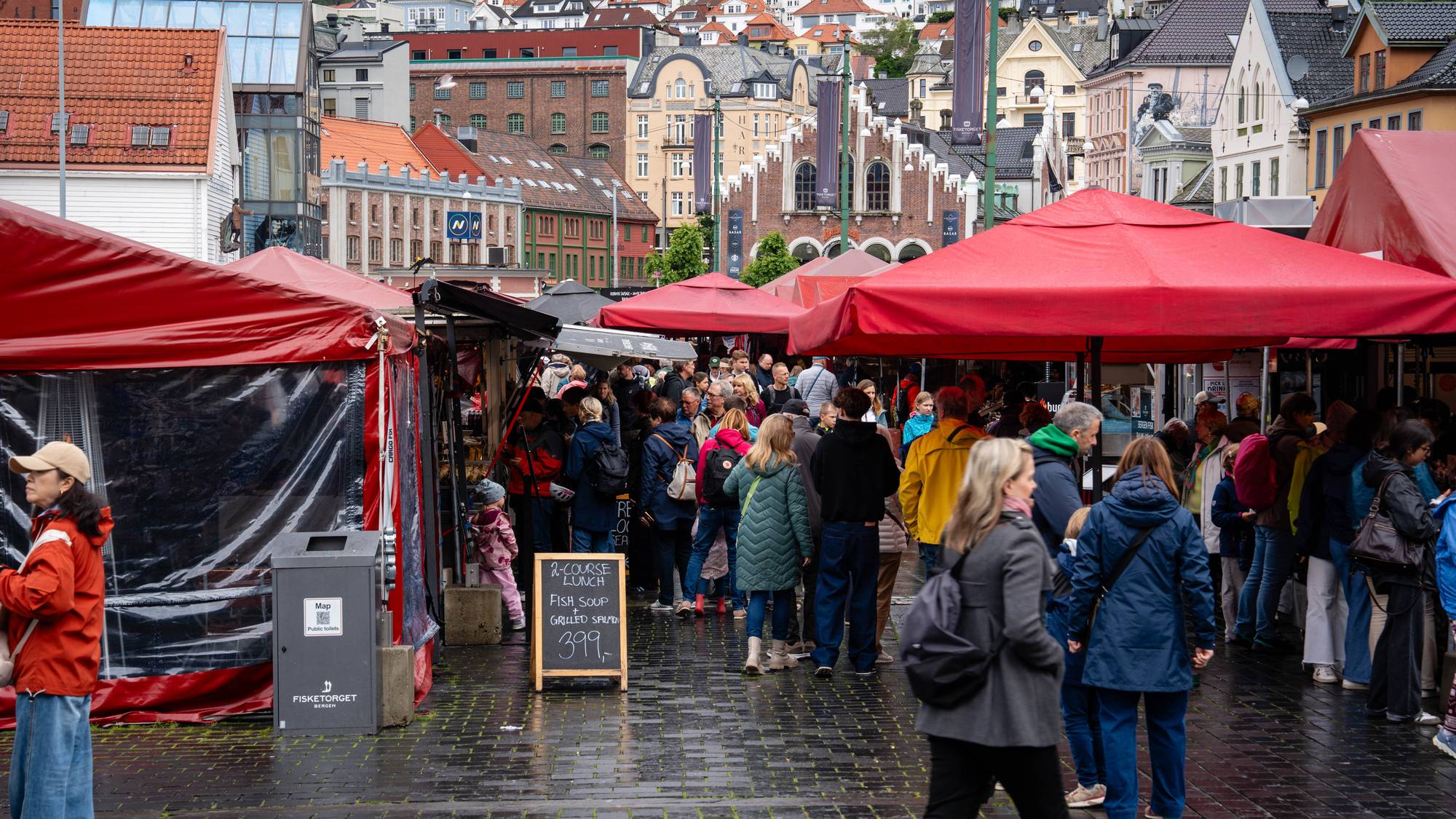 Mennesker går mellom røde markisetelt på Fisketorget i Bergen. Bebyggelse i bakgrunnen. (Bildebeskrivelsen er laget av en KI-tjeneste)
