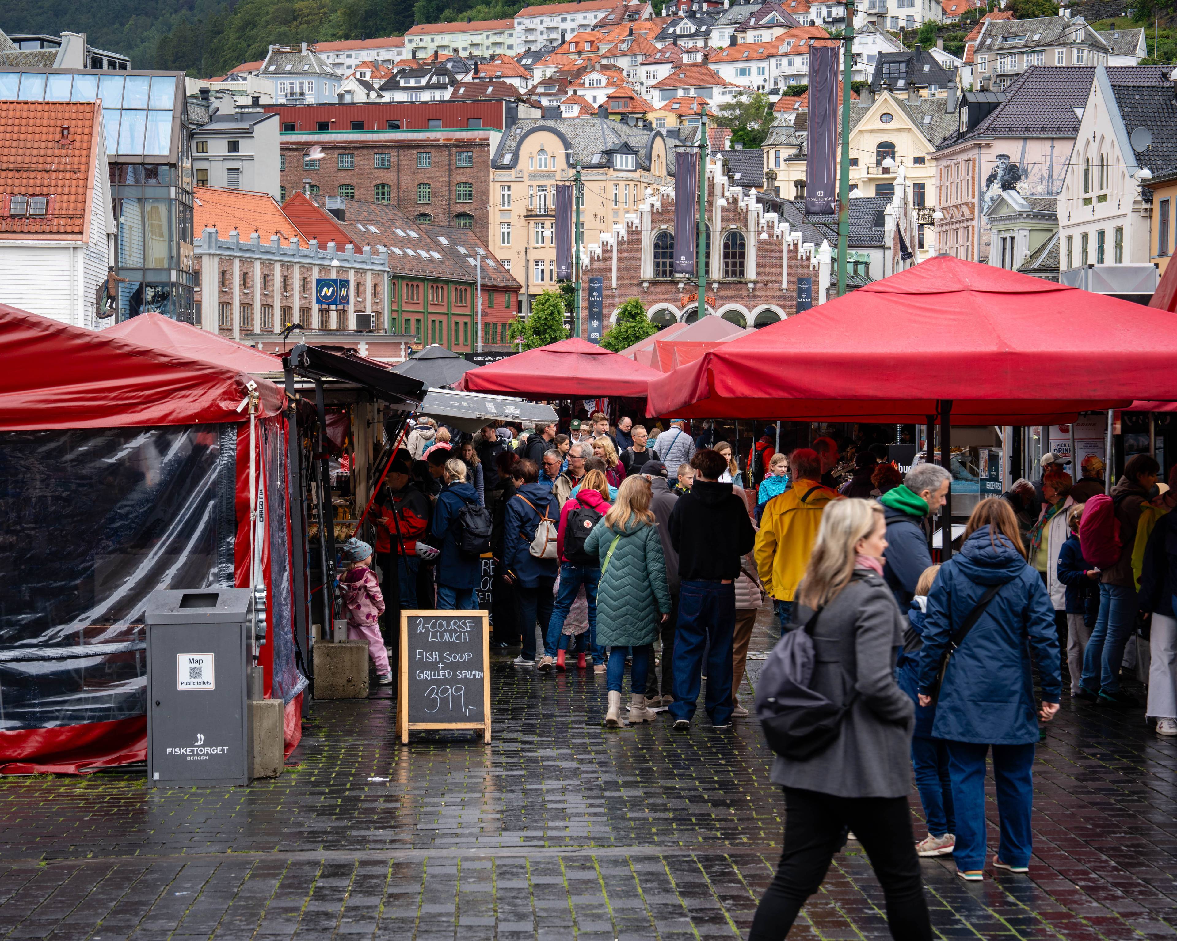 Mennesker går mellom røde markisetelt på Fisketorget i Bergen. Bebyggelse i bakgrunnen. (Bildebeskrivelsen er laget av en KI-tjeneste)