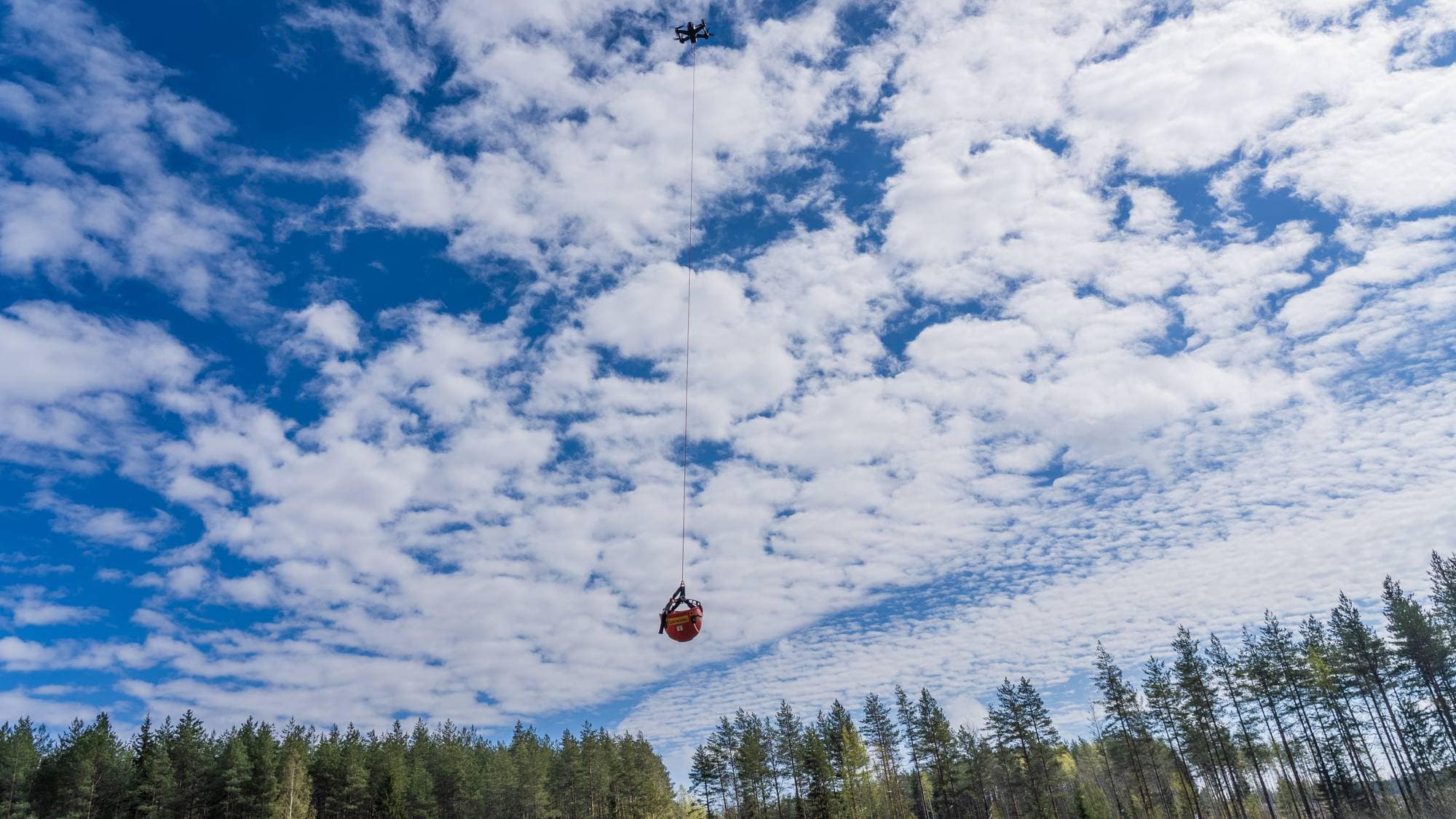 En drone med en vannsekk henger fra et tau mot en blå himmel med skyer. Vannsekken er rød og har et gult symbol. Skog er synlig i bakgrunnen. (Bildebeskrivelsen er laget av en KI-tjeneste)