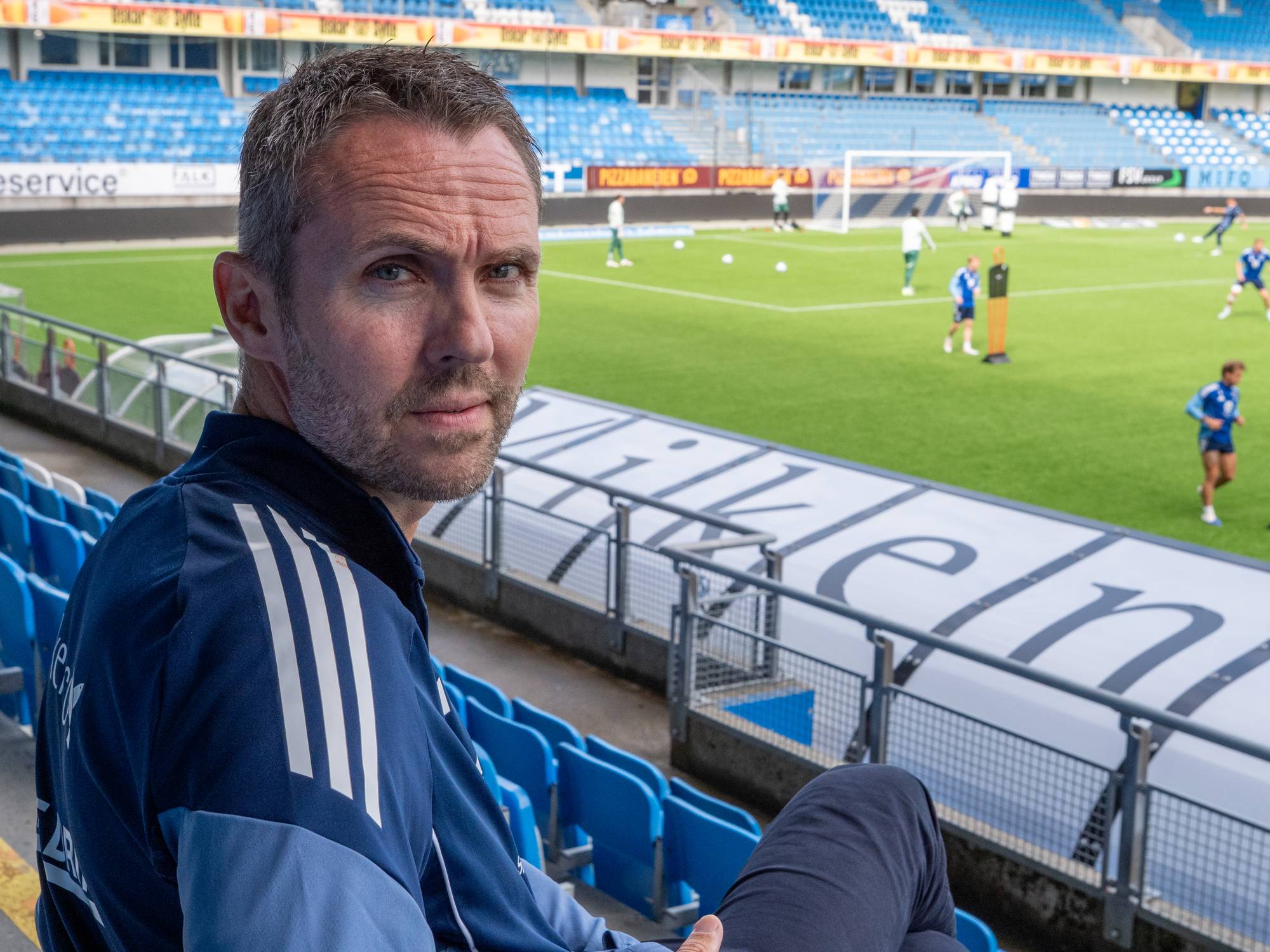 en mann som sitter på en tribune på en fotballbane, på gressmatta har et lag trening 