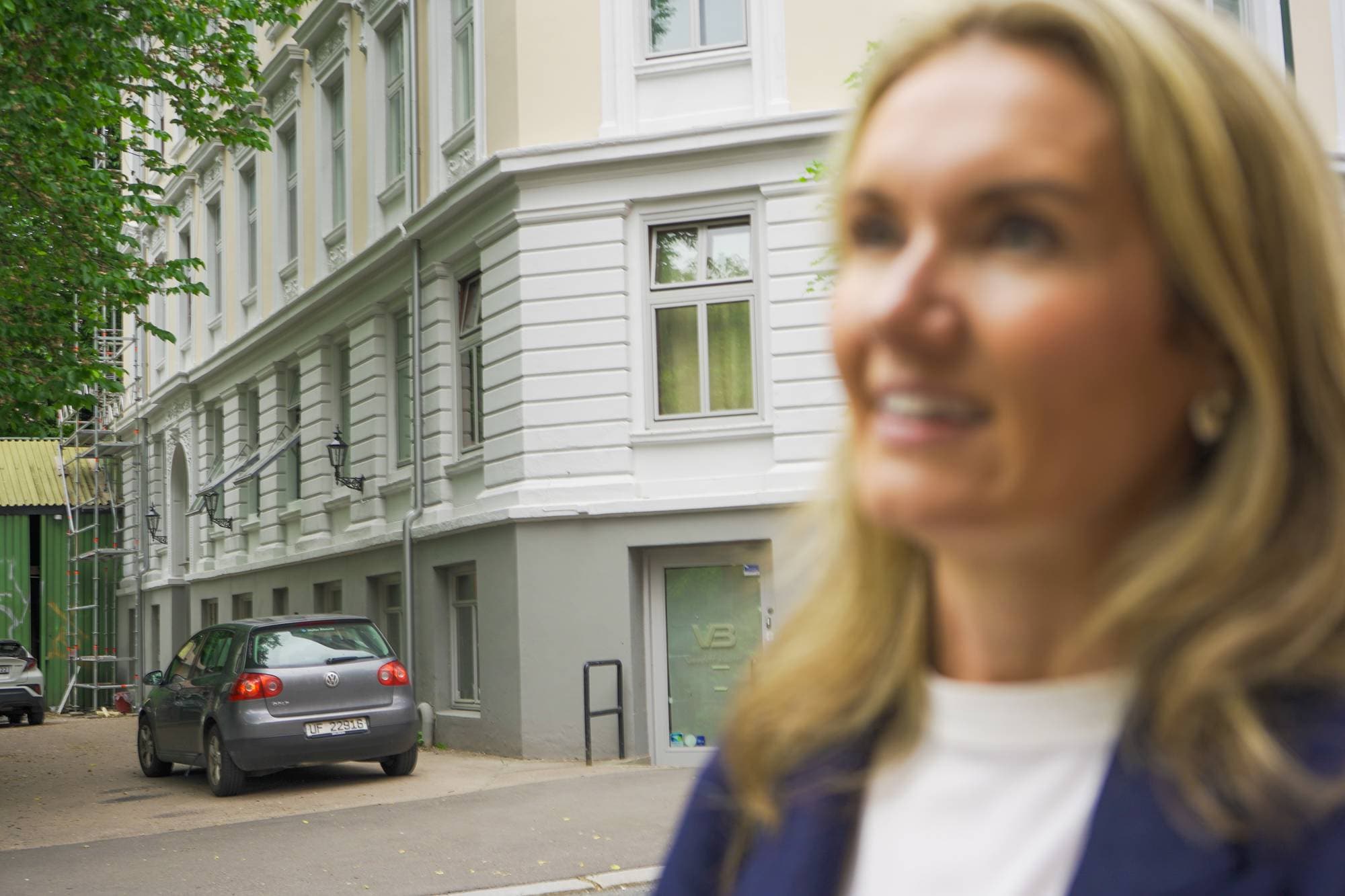 En uskarpt kvinne i forgrunnen smiler til siden. I bakgrunnen er en grå bil parkert utenfor en hvit bygning. Bygningen har tradisjonelle vinduer og detaljer. Utenfor bygningen er det et gjerde og stillaser. (Bildebeskrivelsen er laget av en KI-tjeneste)