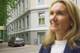 Julianne Kyoo-Ree Eikrem / NRK En uskarpt kvinne i forgrunnen smiler til siden. I bakgrunnen er en grå bil parkert utenfor en hvit bygning. Bygningen har tradisjonelle vinduer og detaljer. Utenfor bygningen er det et gjerde og stillaser. (Bildebeskrivelsen er laget av en KI-tjeneste)