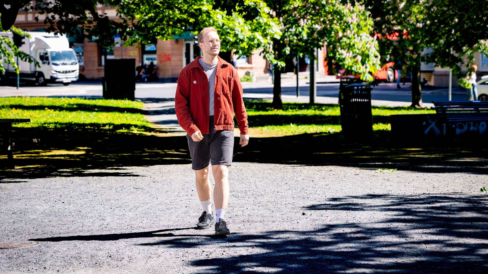 Mann med oransje skjorte og svart kortbukse går mot kameraet på grusveg i park