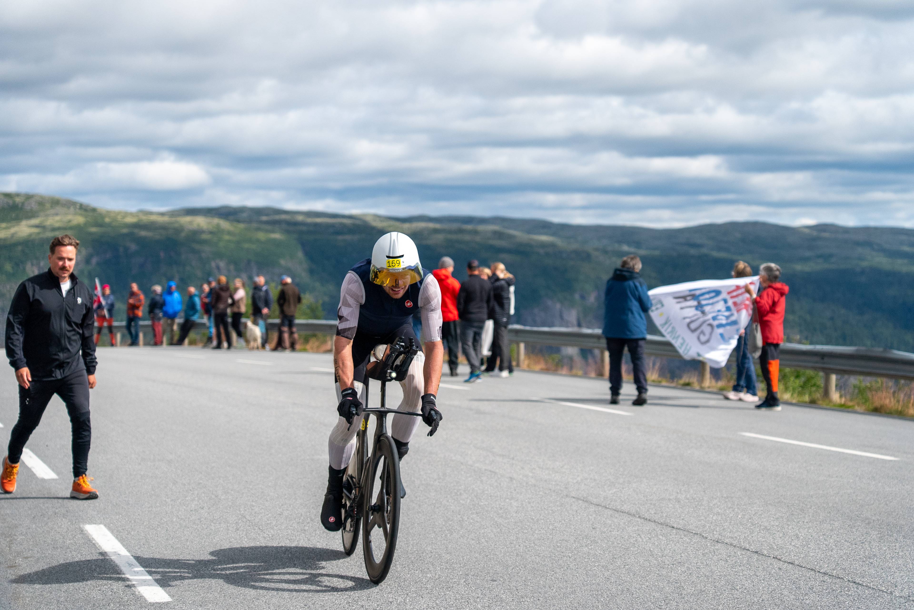 Syklist i tempodrakt sykler på vei i fjellterreng, omgitt av tilskuere. (Bildebeskrivelsen er laget av en KI-tjeneste)