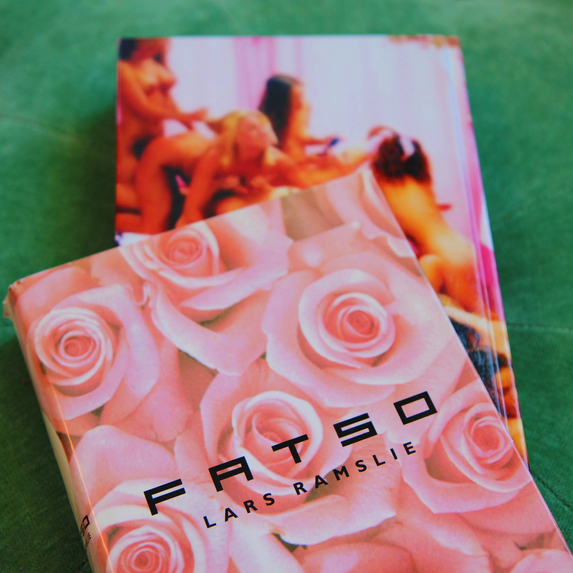 En bok ligger på en grønn fløyelssofa, papiromslaget er delvis av. Den har et design med rosa roser og teksten "FATSO LARS RAMSLIE". En del av boken skimtes bak. Den har et bilde av nakne damer som gjør seksuelle tung med hverandre.