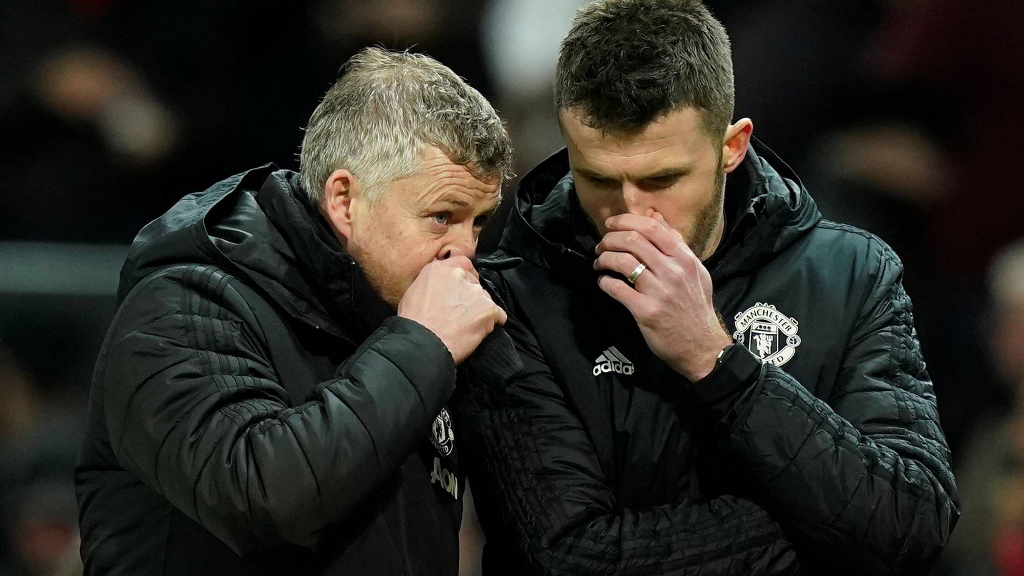 To menn i svarte jakker med Manchester United-logo snakker tett sammen. Den ene mannen lener seg inn mot den andre og begge har hendene foran munnen. Bildet er tatt på en mørk arena med publikum i bakgrunnen. (Bildebeskrivelsen er laget av en KI-tjeneste)