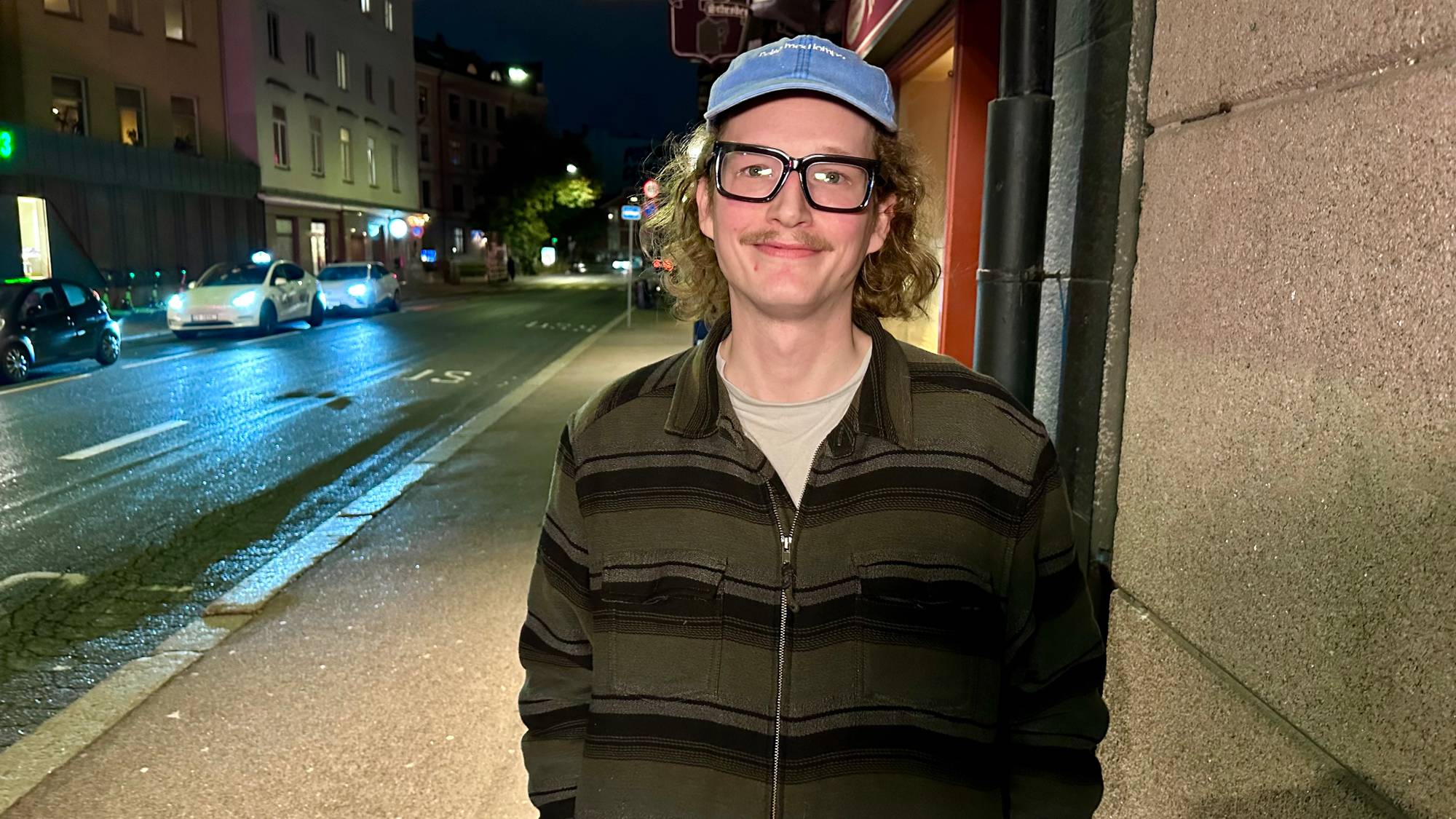 En mann med blå caps og briller står på en fortau i en bygate om kvelden. (Bildebeskrivelsen er laget av en KI-tjeneste)