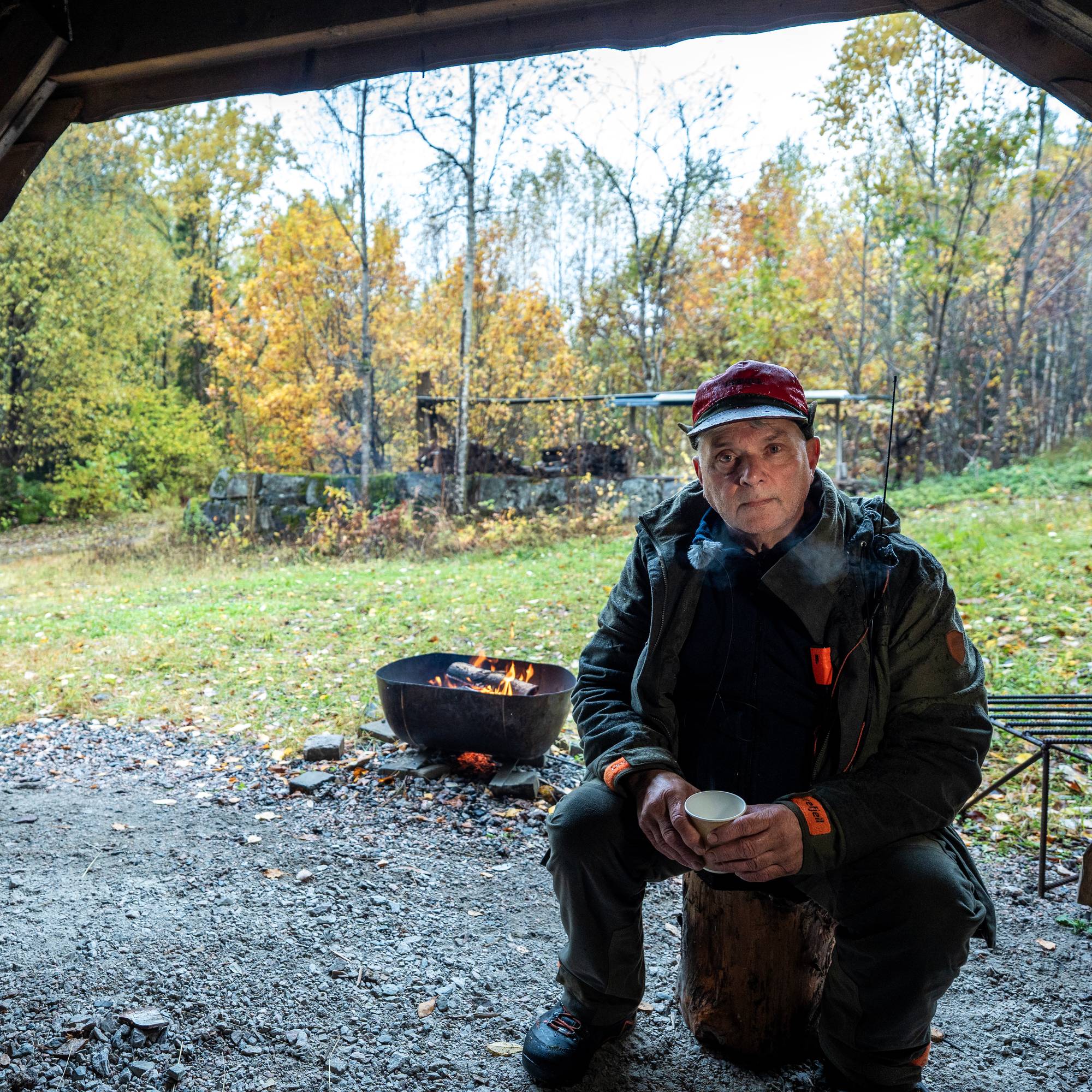 En mann i jaktklær sitter på en trestokk foran et bål i et gapahuk i skogen. (Bildebeskrivelsen er laget av en KI-tjeneste)