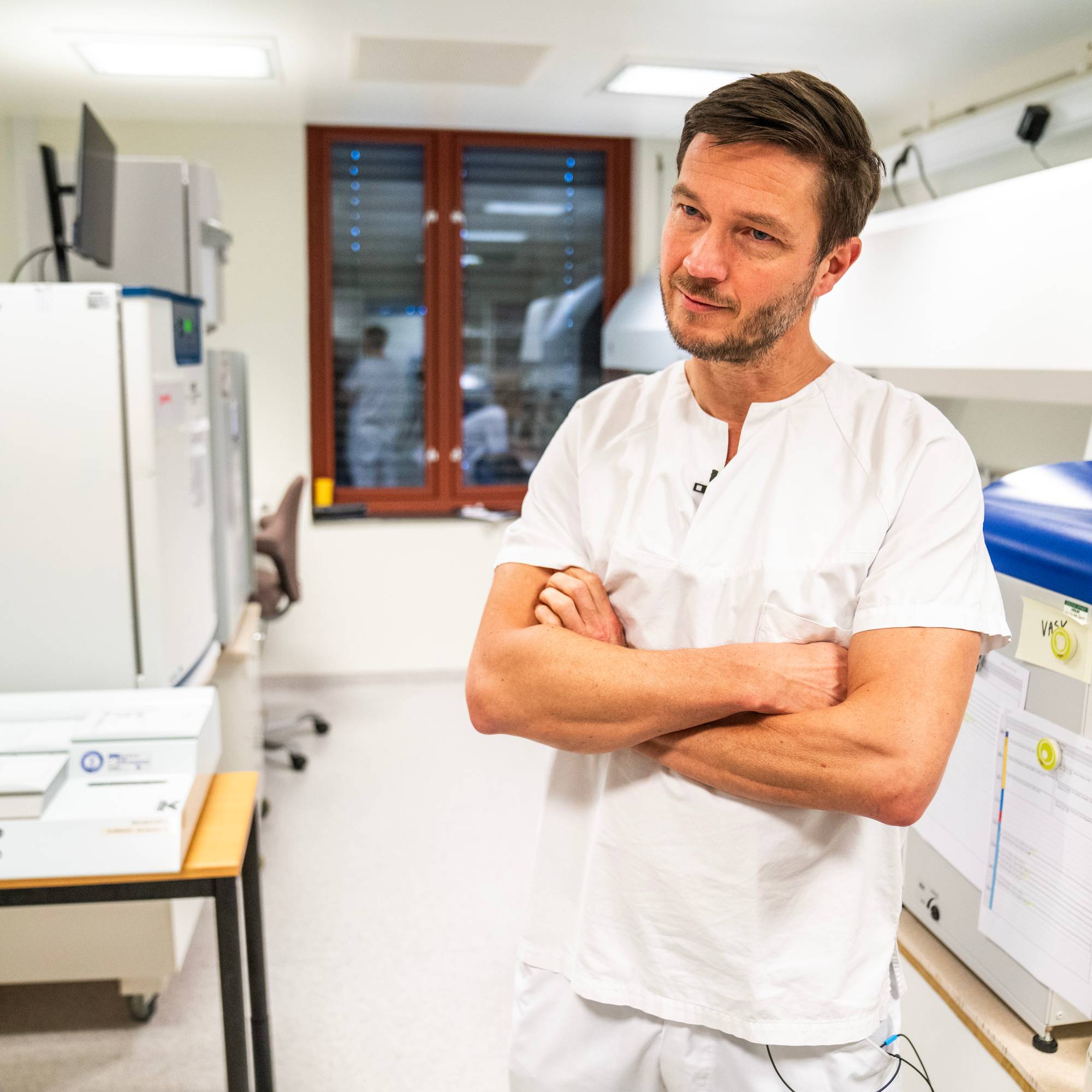 En lege i hvit uniform står med hendene i kors i et laboratorium.