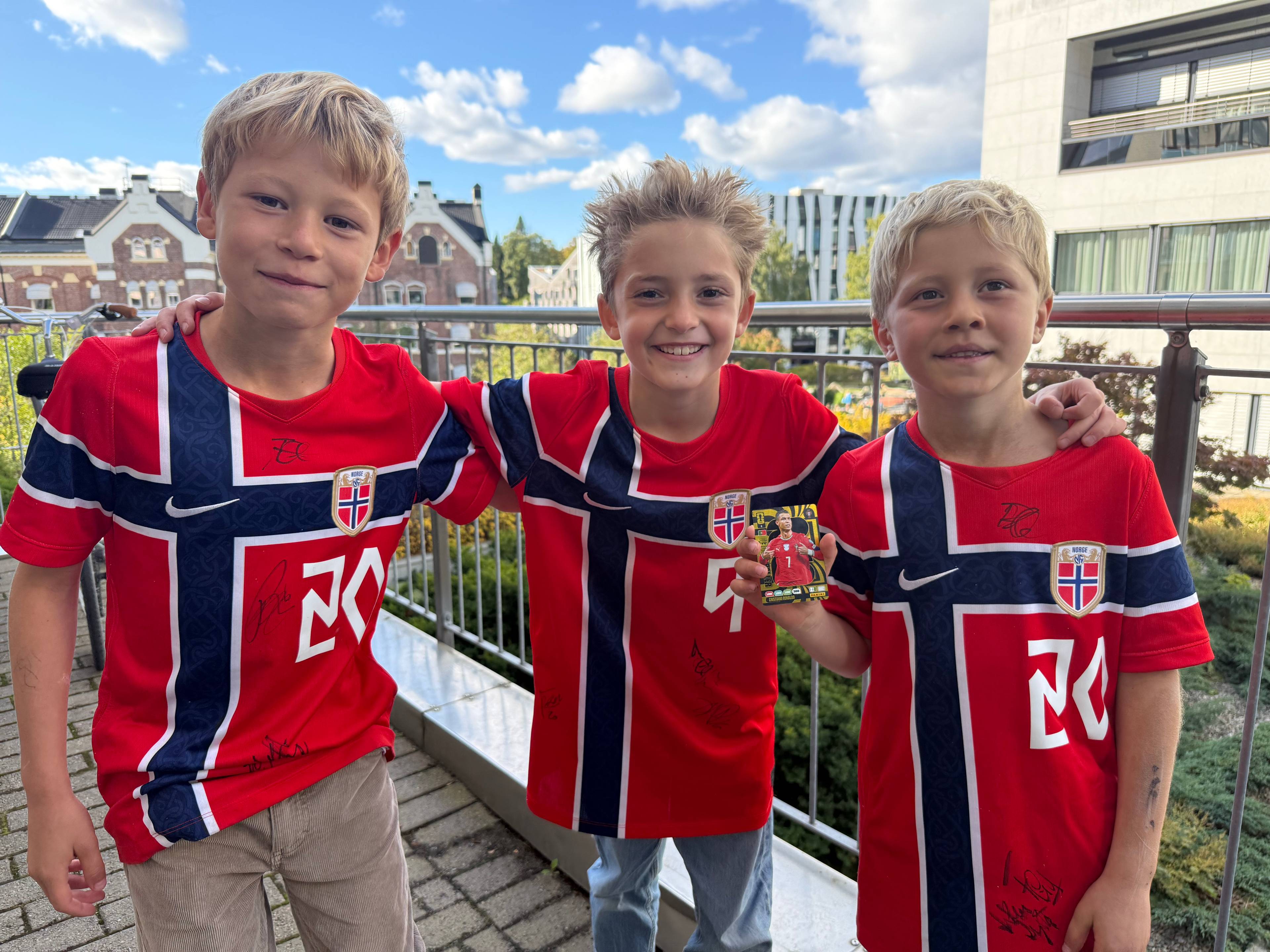 Tre gutter i norske landslagsdrakter står sammen utendørs. En av dem holder et fotballkort. (Bildebeskrivelsen er laget av en KI-tjeneste)