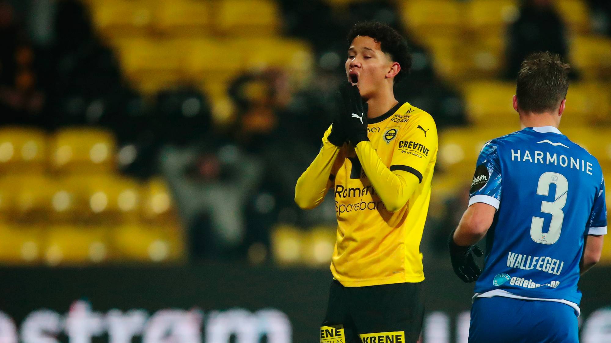 En fotballspiller i gul drakt med hendene foran munnen ser til venstre. Bak ham står en spiller i blå drakt. Publikum sitter på tribuner i bakgrunnen. Et reklameskilt med tekst er synlig nederst på bildet. (Bildebeskrivelsen er laget av en KI-tjeneste)