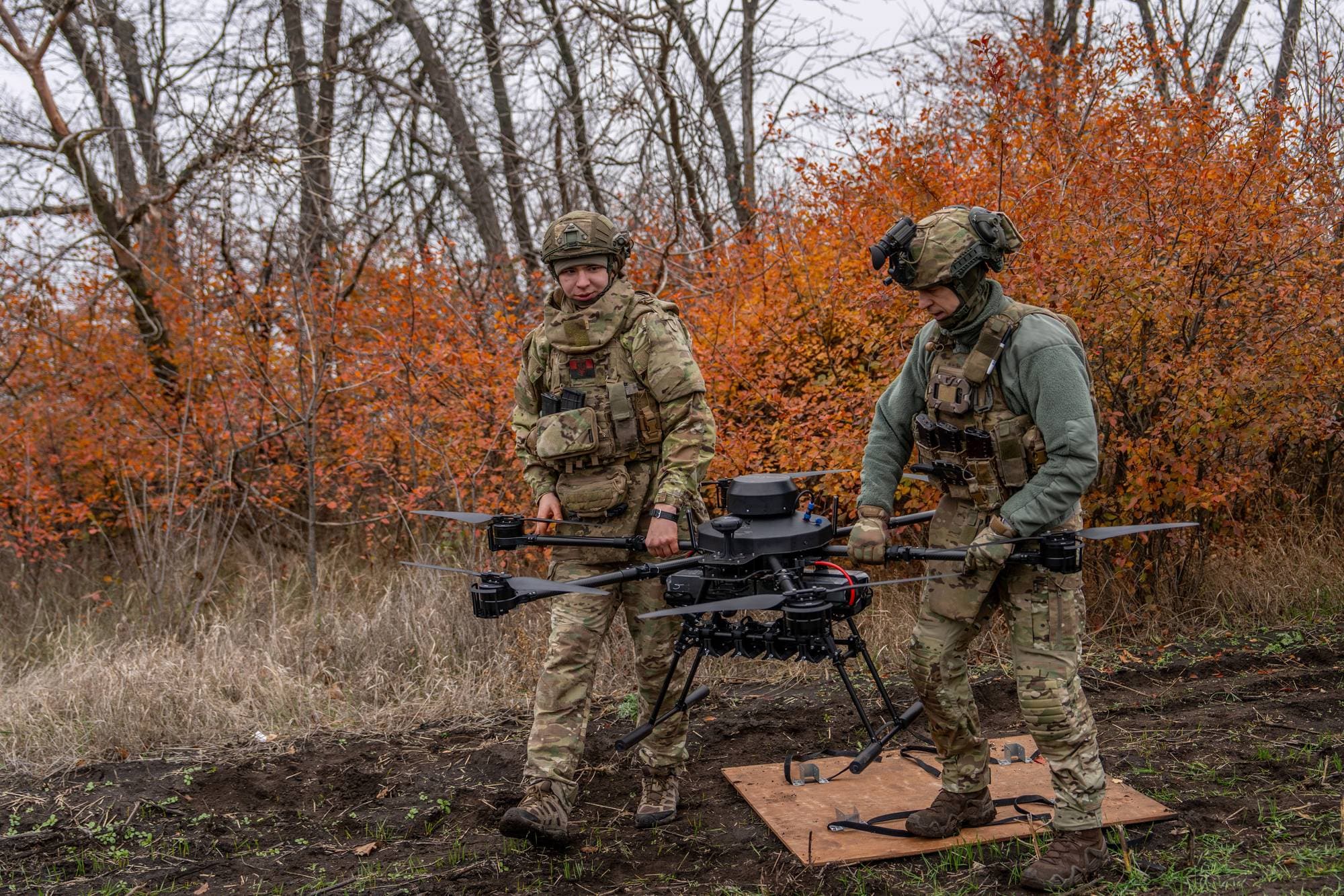 To soldater i militæruniform holder en stor drone i et utendørsområde. Dronen har flere propeller og en kameraboks. Soldatene er utstyrt med hjelmer, taktiske vester og annet militært utstyr. Vegetasjonen rundt dem er preget av høstfarger. (Bildebeskrivelsen er laget av en KI-tjeneste)