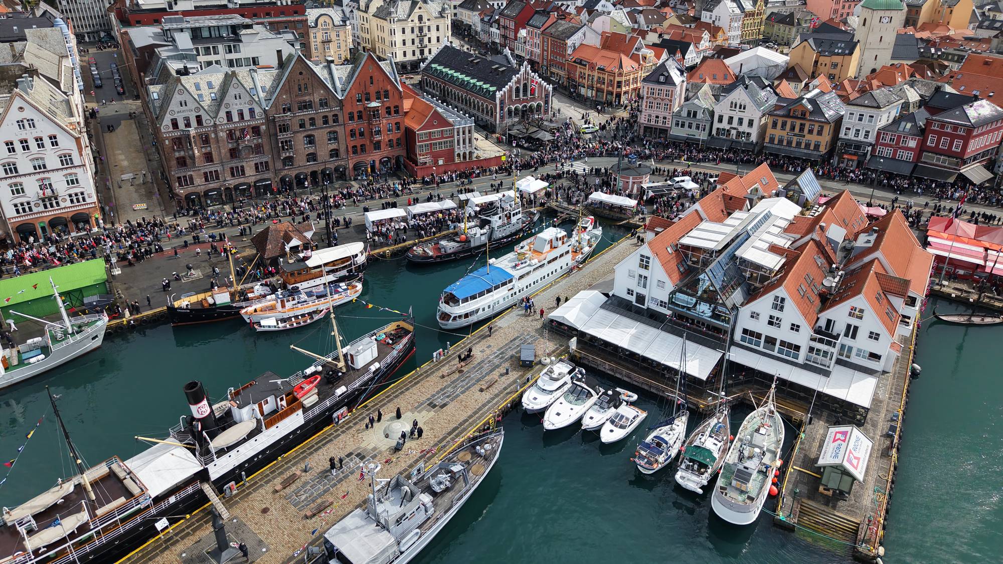 Dronefoto som viser områder av Bryggen, Zachariasbryggen og Torget i Bergen sentrum. Store folkemengder var samlet i sentrum for å se på prosesjonen og feire nasjonaldagen. 