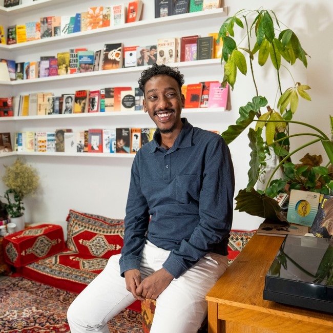 Forfatter Hamza Hirsi sitter på en puff ved siden av et skrivebord. Bak ham er veggene dekket av fargerike bøker. En arabisk sofa og et fargerikt teppe dekker gulvet.