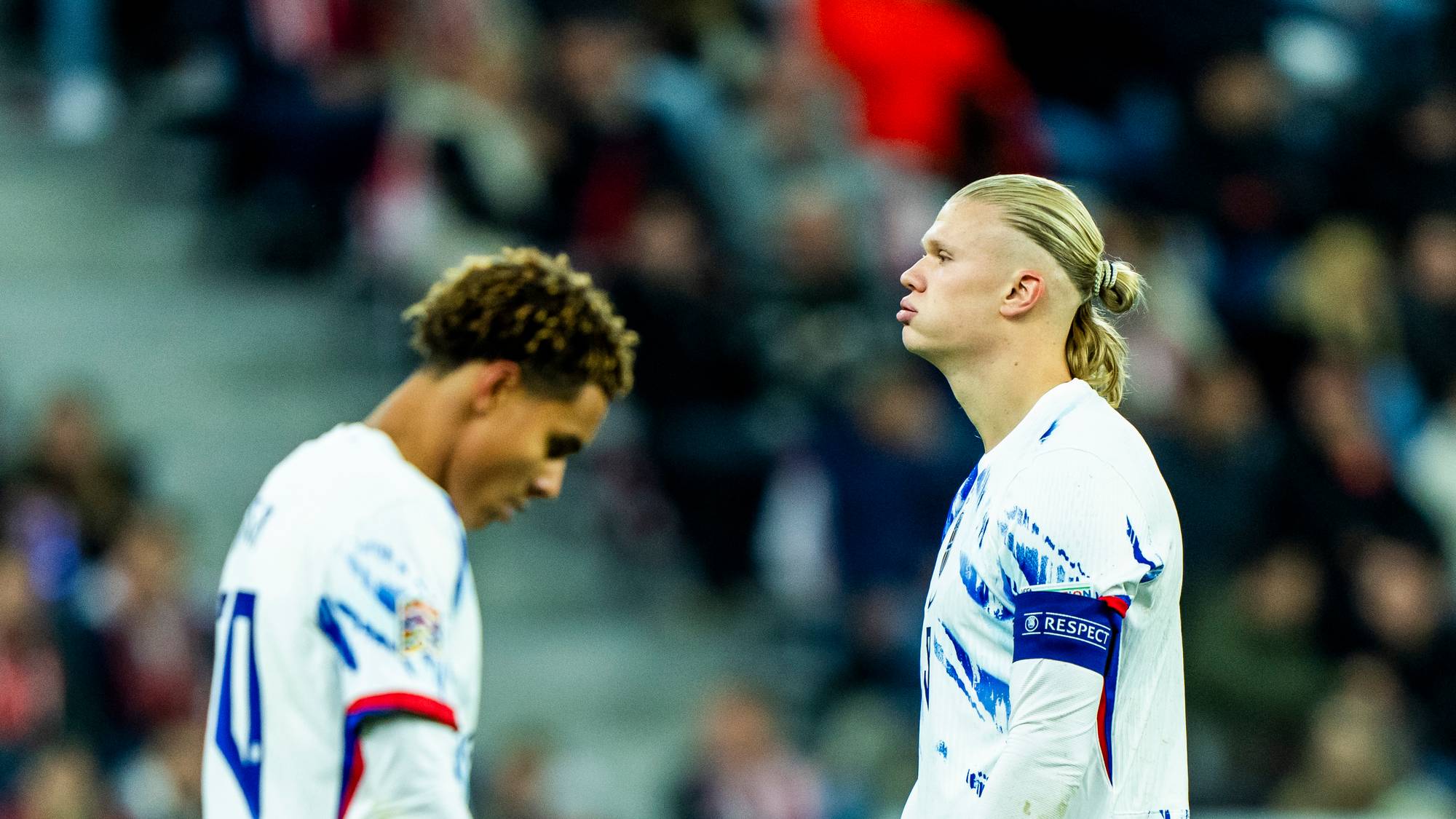 Erling Braut Haaland og en lagkamerat i hvite drakter på en fotballbane med publikum i bakgrunnen. (Bildebeskrivelsen er laget av en KI-tjeneste)