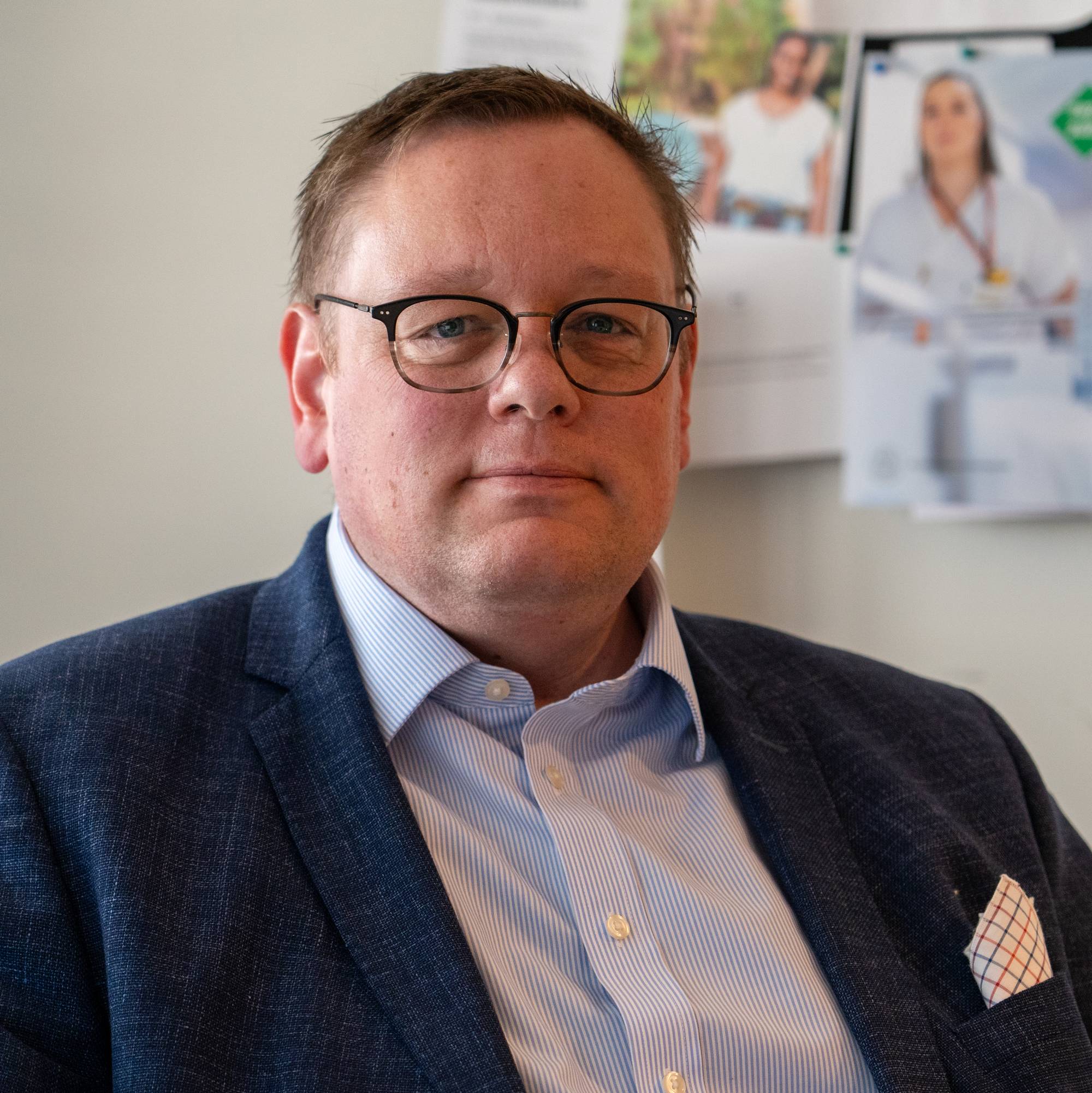 Portrett av en mann med briller, kledd i blå dressjakke og stripete skjorte. Mannen ser mot kamera. Bak ham er det et oppslag med bilder og tekst, derav ett med en sykepleier. (Bildebeskrivelsen er laget av en KI-tjeneste)