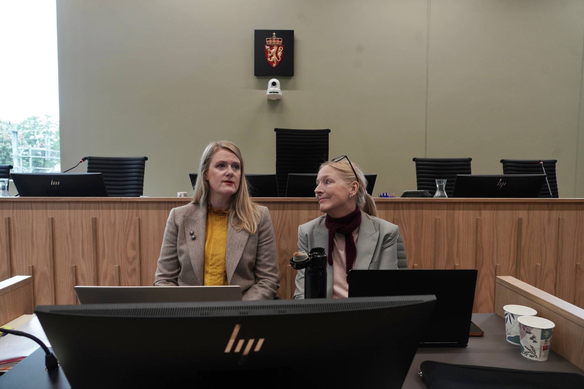 To kvinner i lyse blazere sitter ved et bord i en rettssal foran laptoper. Riksvåpenet henger på veggen bak. 